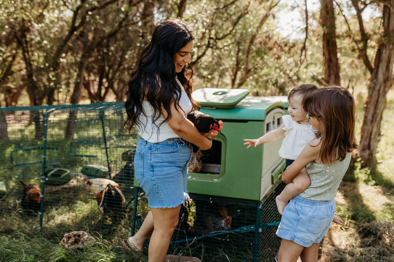 A mother and two young daughters interact with chickens at a green backyard chicken coop in the woods.