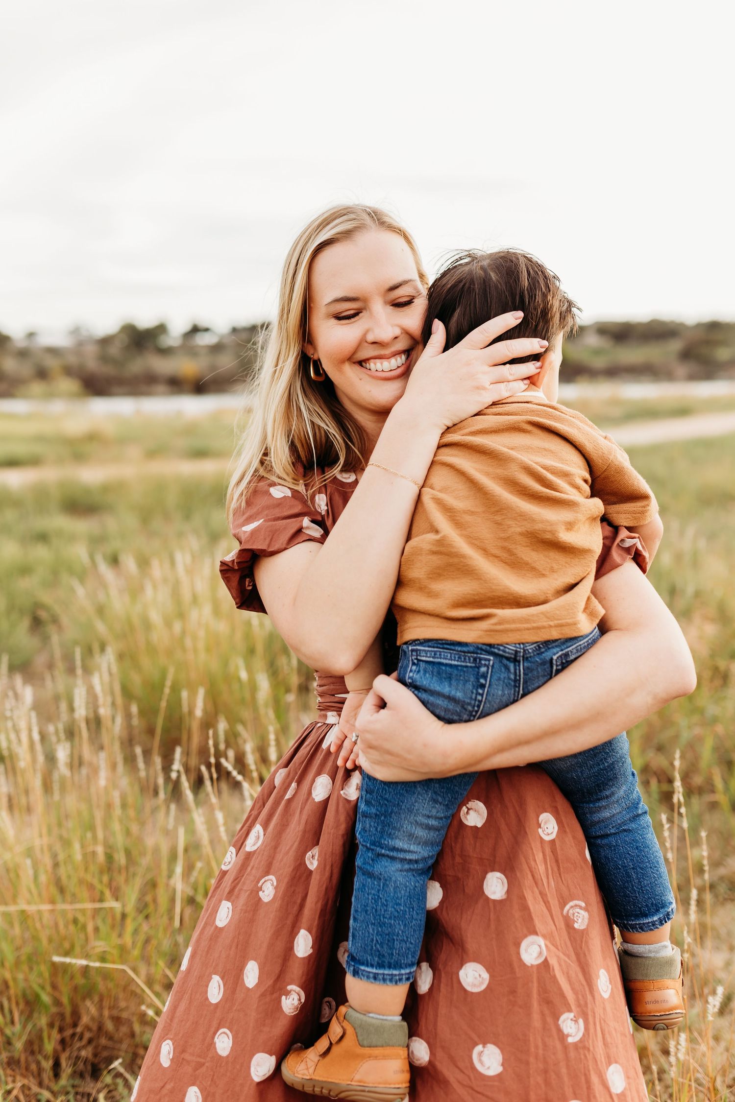 A happy blonde mother in a brown polka dot dress hugs her toddler son tightly while standing in a grassy field.