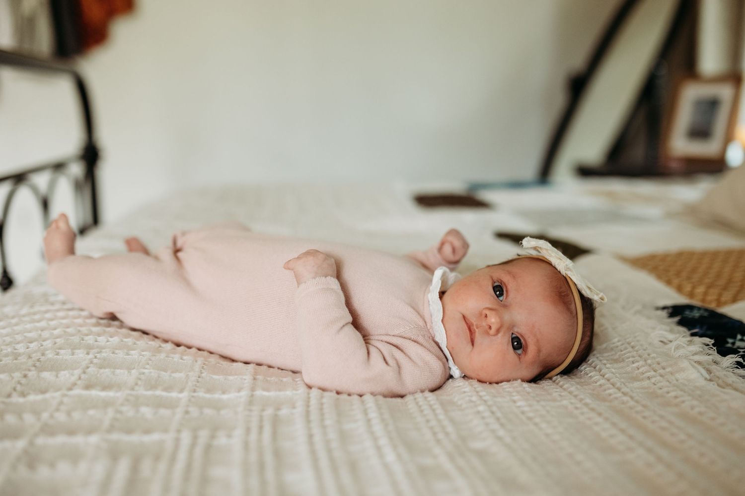A newborn baby girl in a pink onesie with a white bow headband lies peacefully on a white quilted bed.
