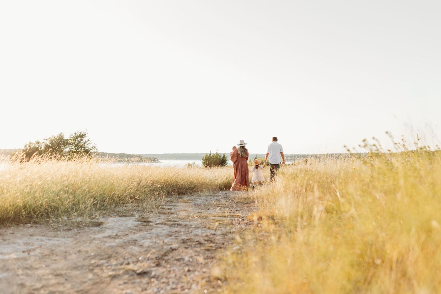 Family walking together on sandy path through tall golden grass toward shimmering lake.