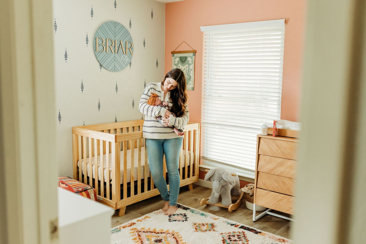 A mother holds her newborn baby in a peachy pink nursery with a wooden crib.