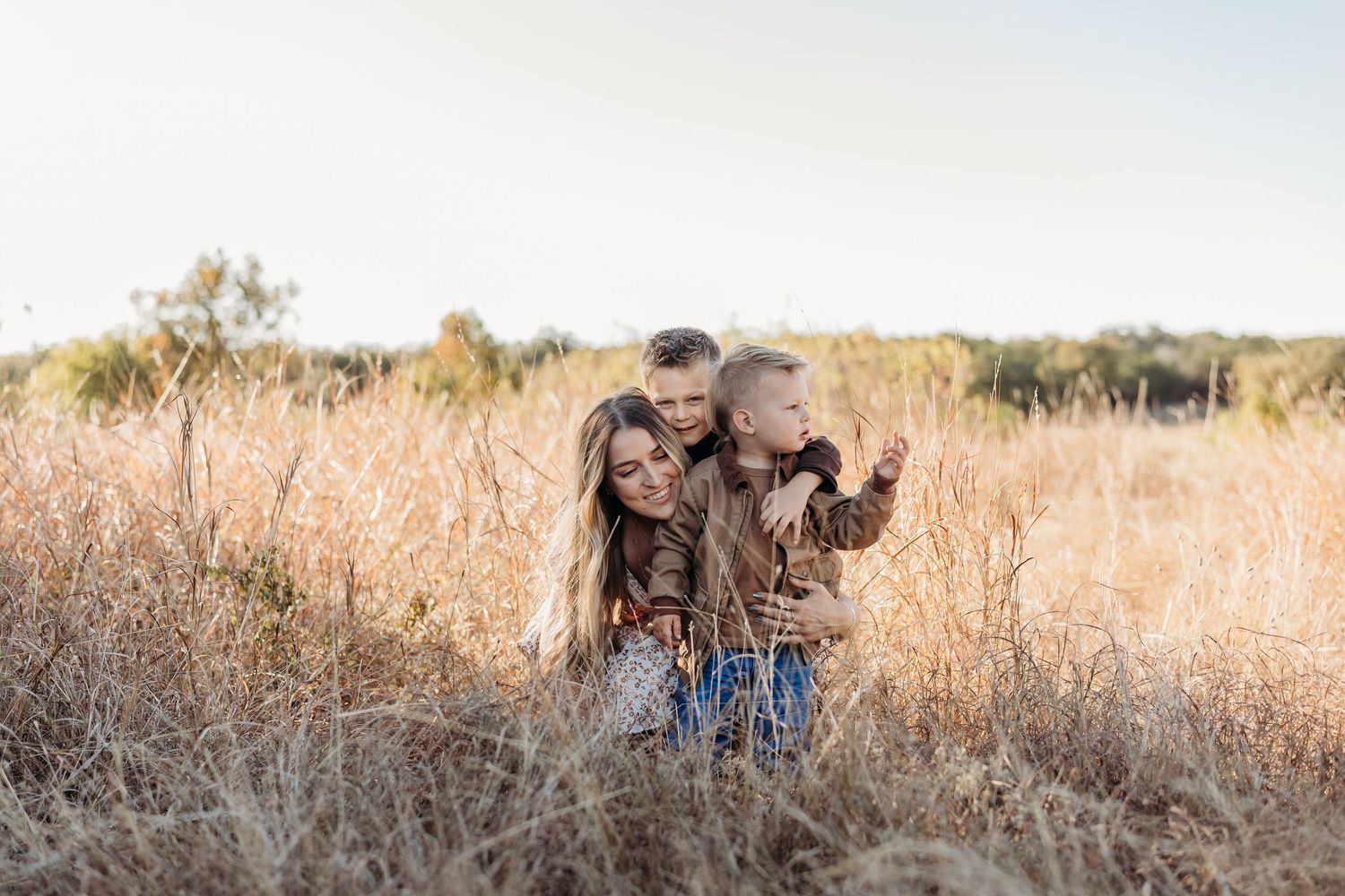 Mother embracing two young boys in a sun-drenched field of tall golden grass.