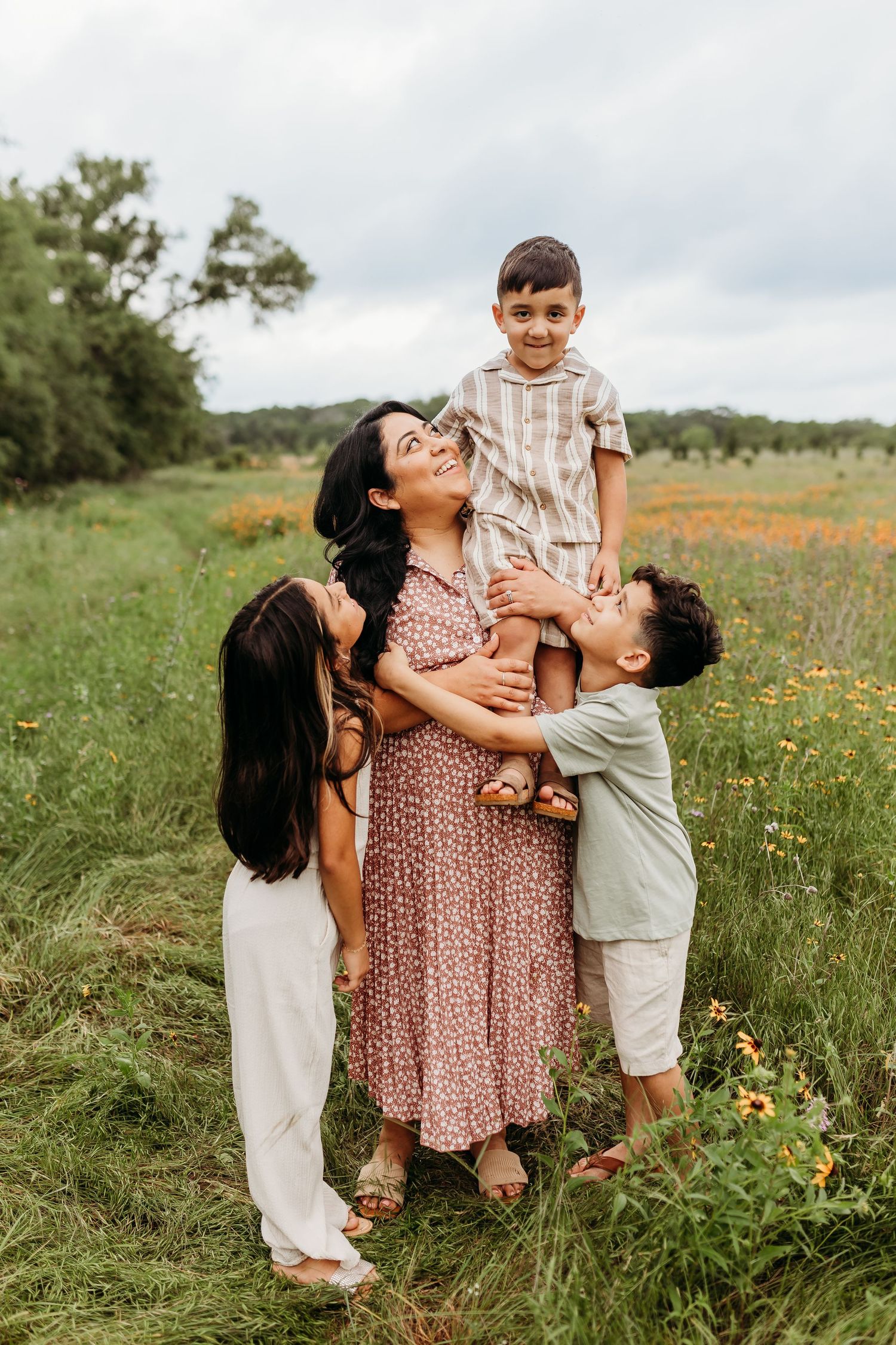 A mother holds her son while her two children embrace her in a colorful wildflower meadow.