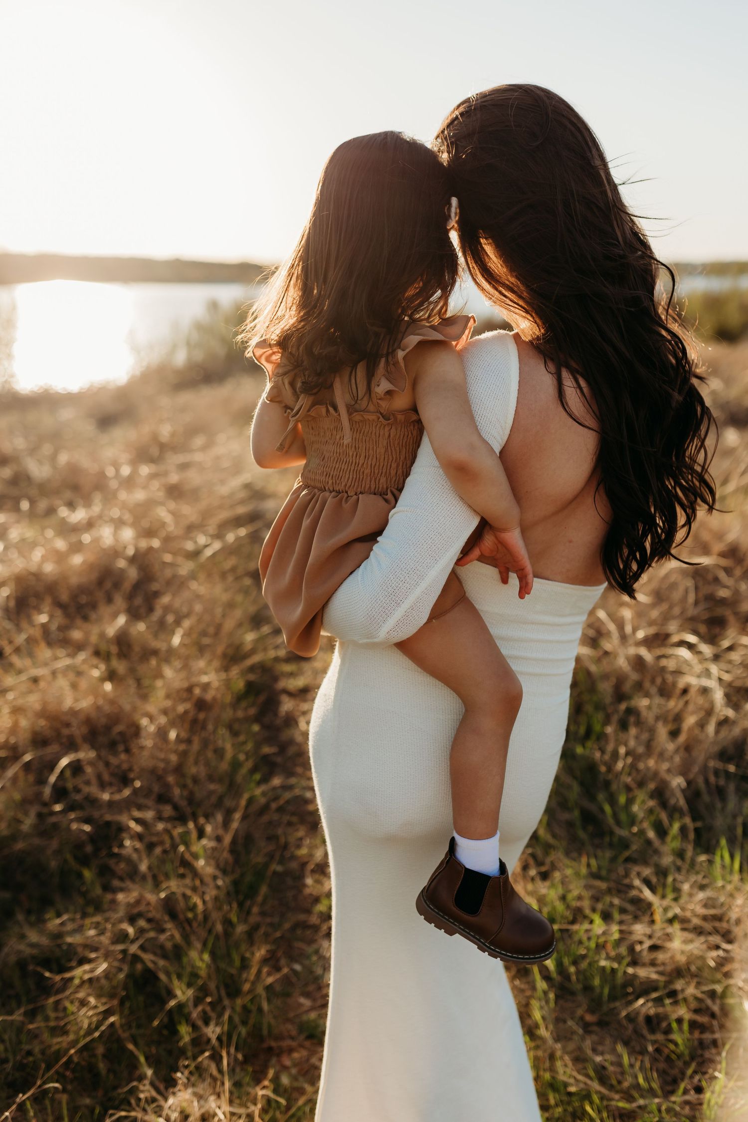 Dark-haired pregnant woman in white dress kissing her toddler daughter in a golden sunset field.