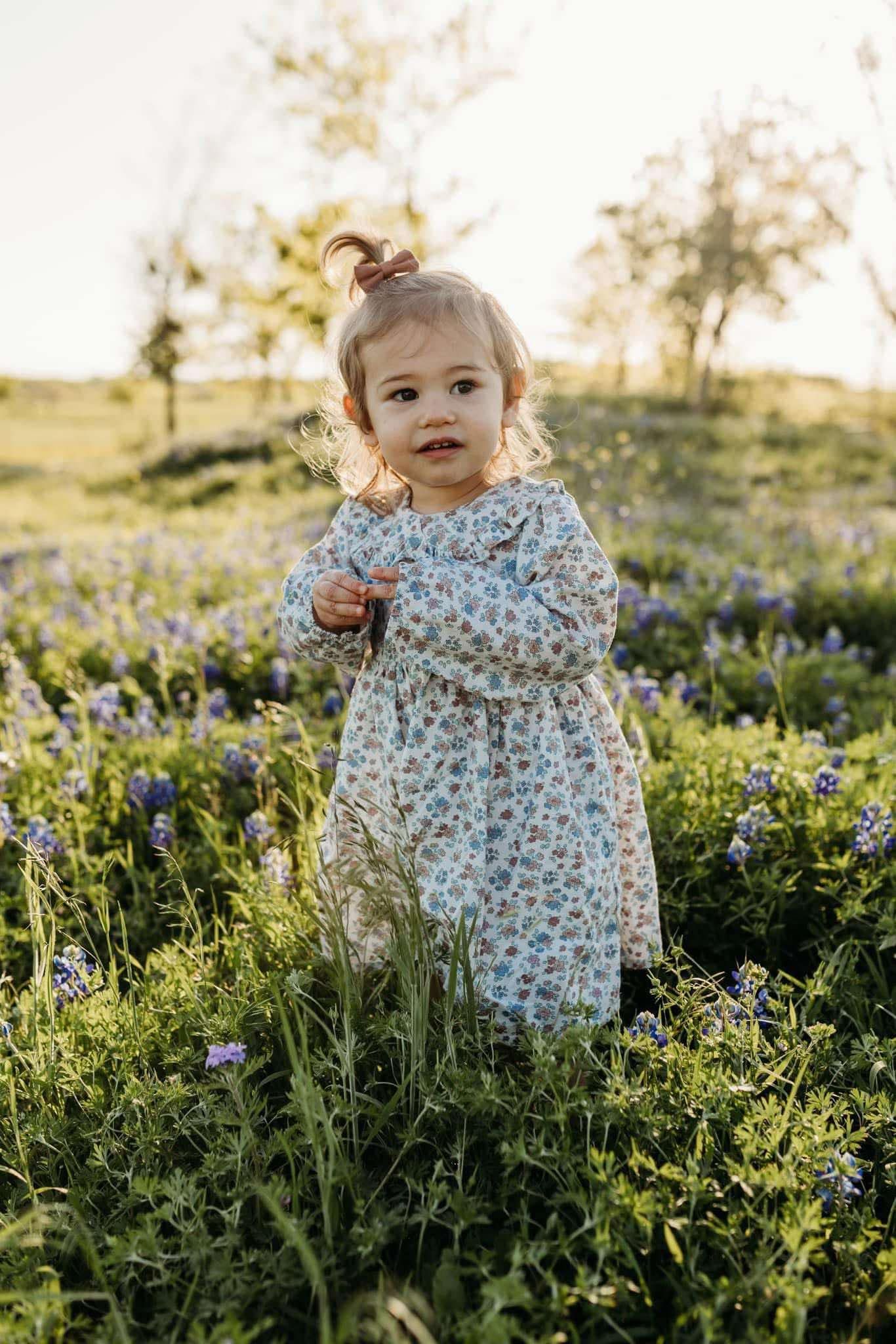 A toddler girl in a floral dress stands in a sunlit field of bluebonnets.