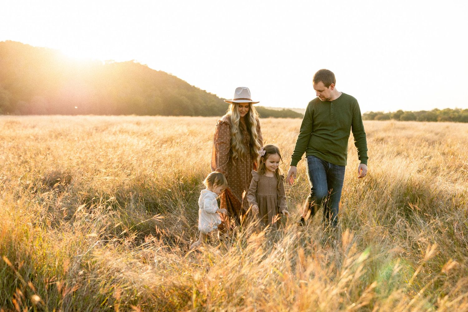 Family of four walking through golden sunlit field at sunset with rolling hills behind them.