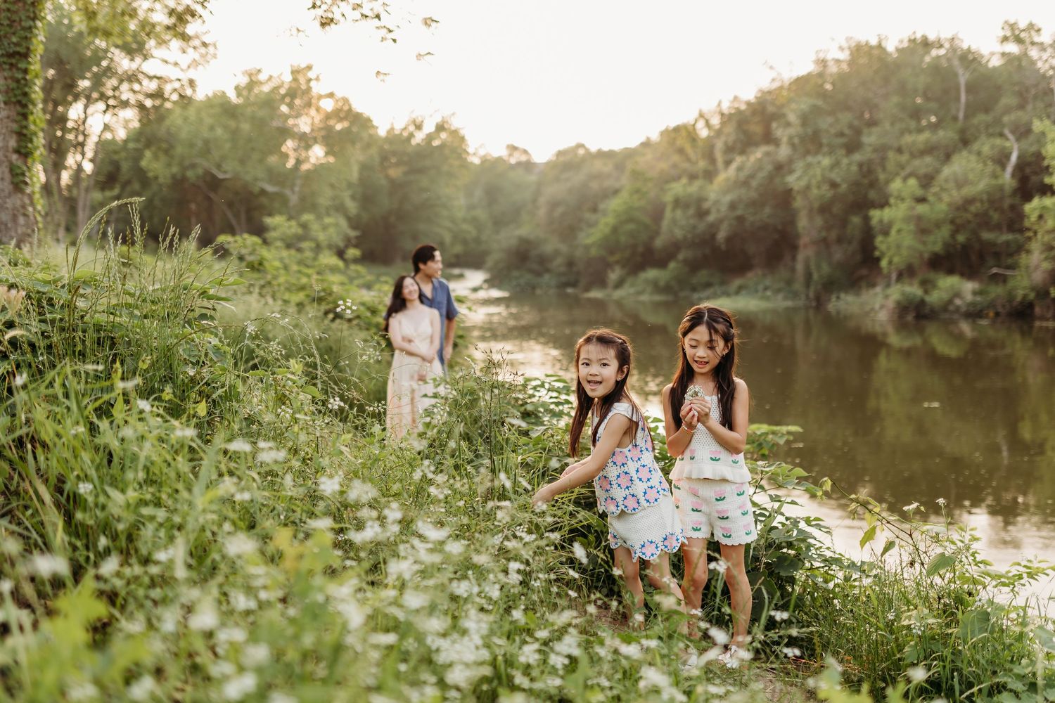 Two young girls play near a wildflower-lined riverbank while their mother stands nearby in golden hour light.