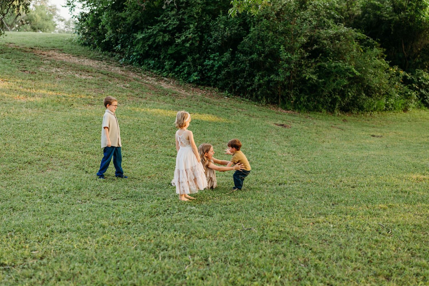 Three children playing together on a green grassy hill surrounded by lush trees outdoors.