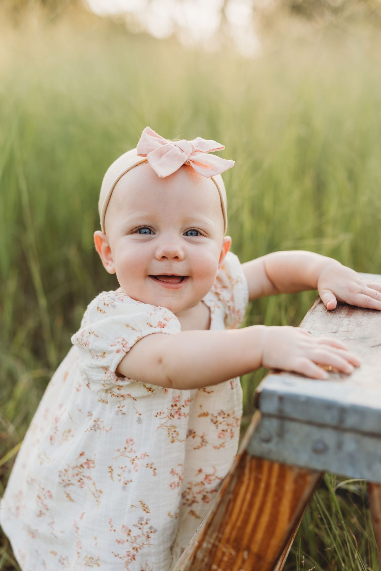 Smiling baby girl in floral dress and pink bow headband standing in green field.