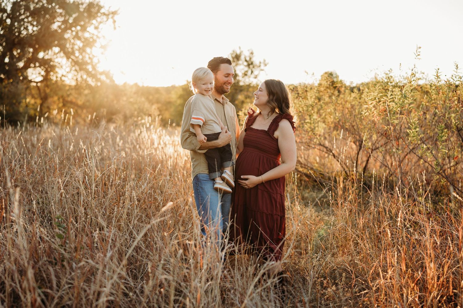 Expecting family with toddler posing in a golden meadow at sunset during maternity photo session.