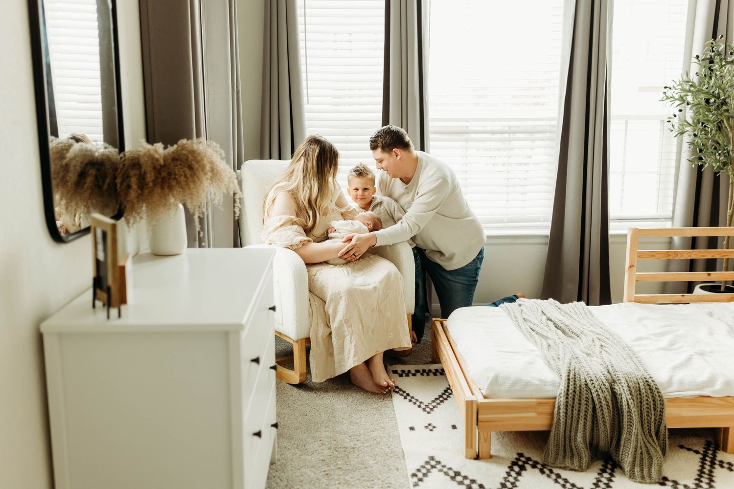 Young family of four sits together in bright bedroom admiring their newborn baby.