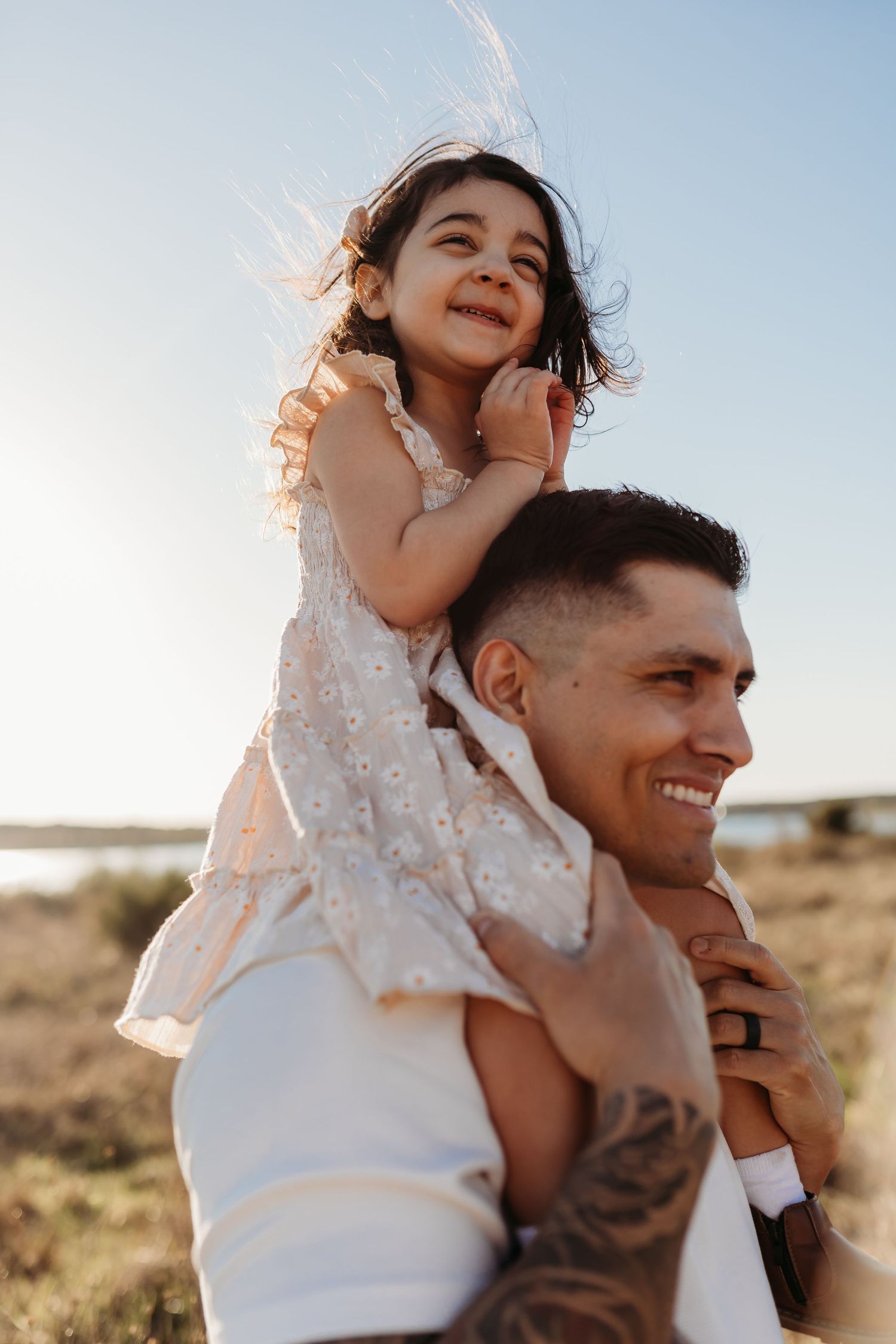 Smiling father with tattoos lifting happy toddler girl on his shoulders against a clear blue sky.