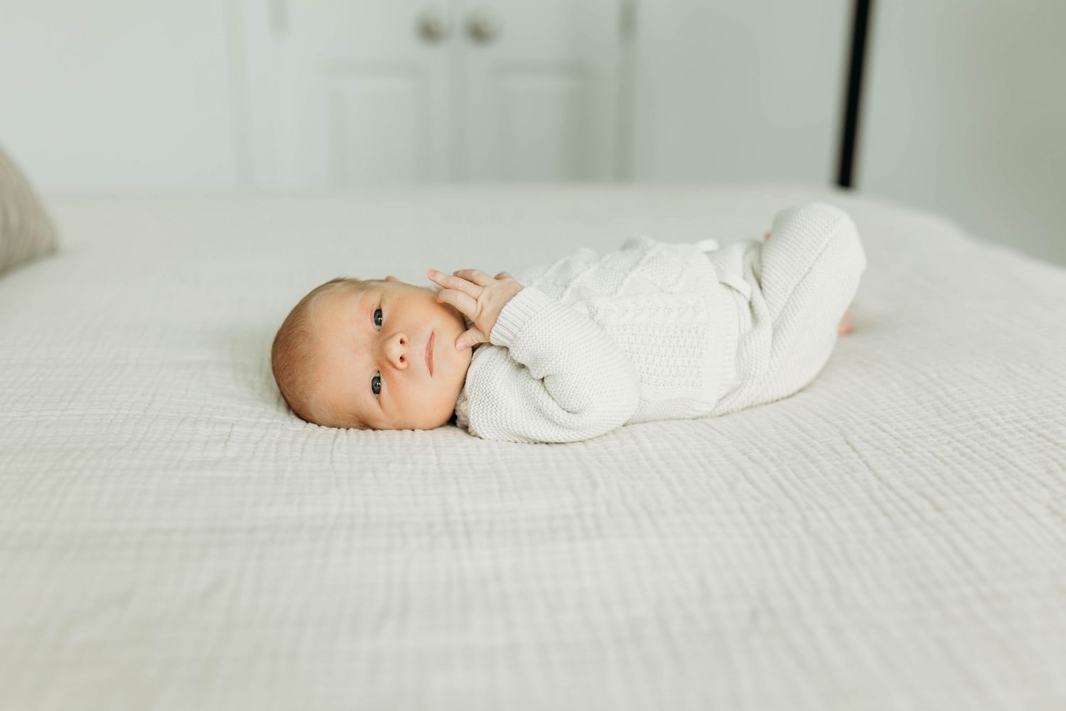 Adorable newborn baby lying on white textured bed, looking up with bright eyes.