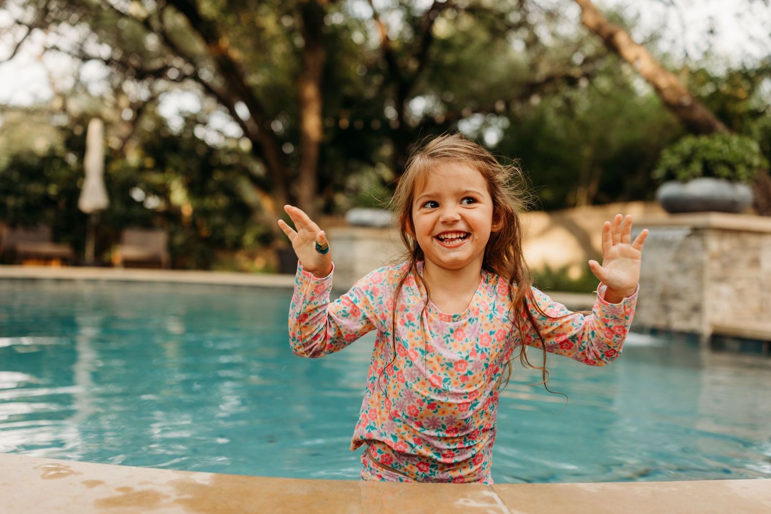 A joyful young girl in a floral swimsuit splashing in a backyard pool with hands raised and a big smile.