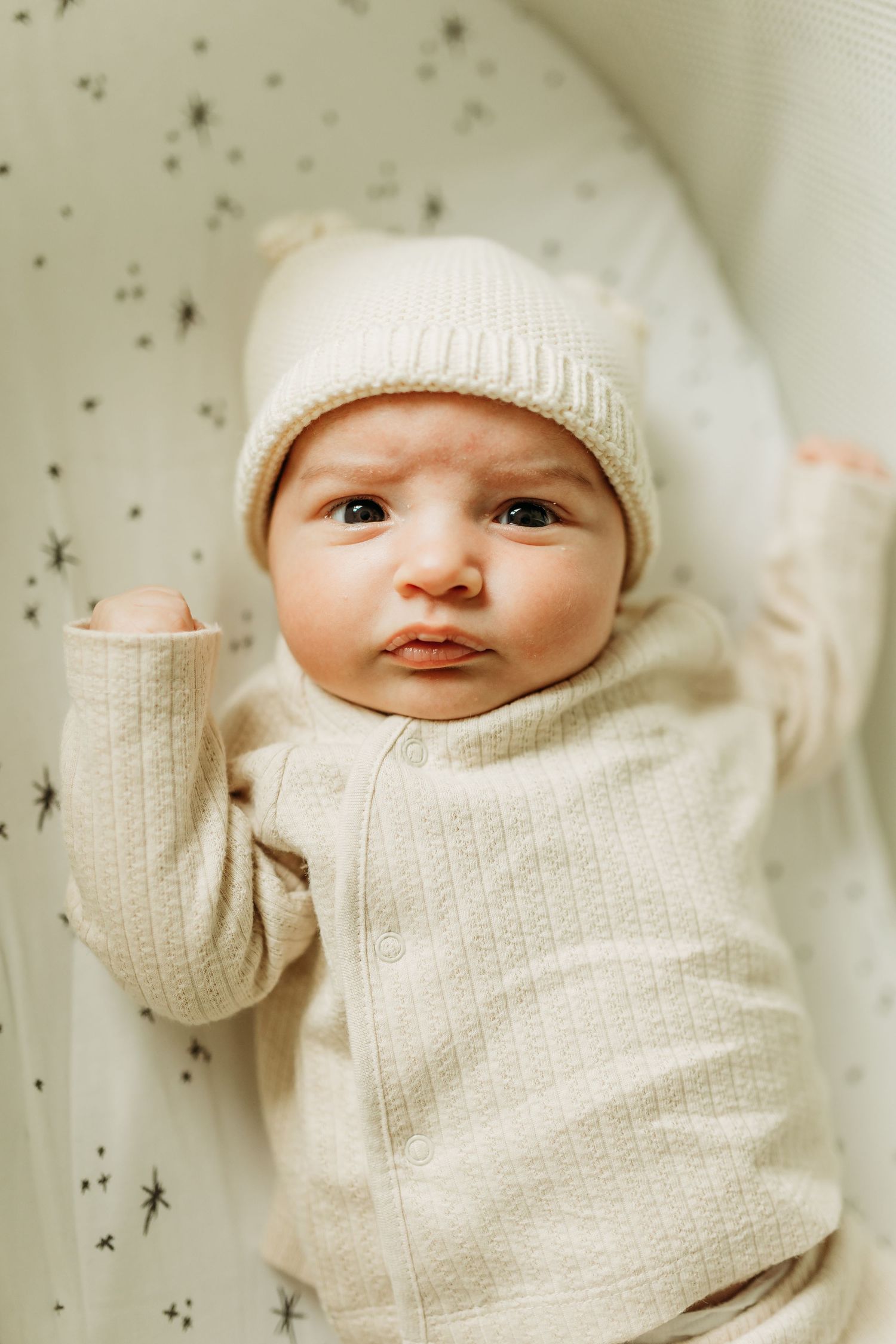 Newborn baby in cream knit bear hat and matching outfit lies in star-print bassinet looking up.