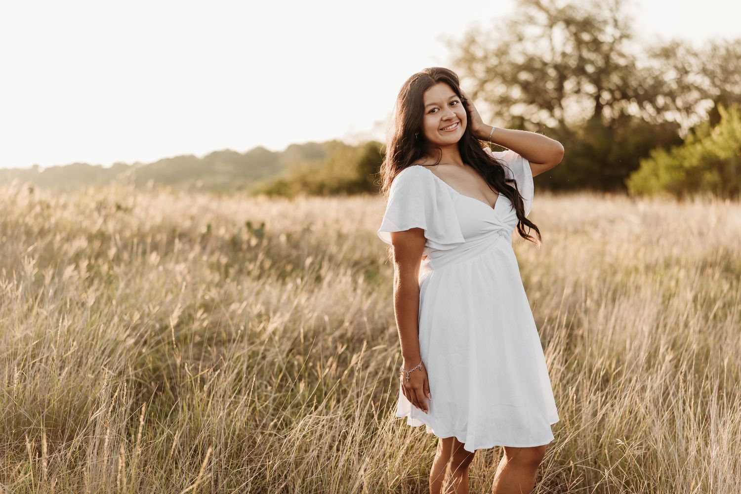 Smiling young woman in a white dress posing in a sunlit golden grass field during outdoor portrait session.