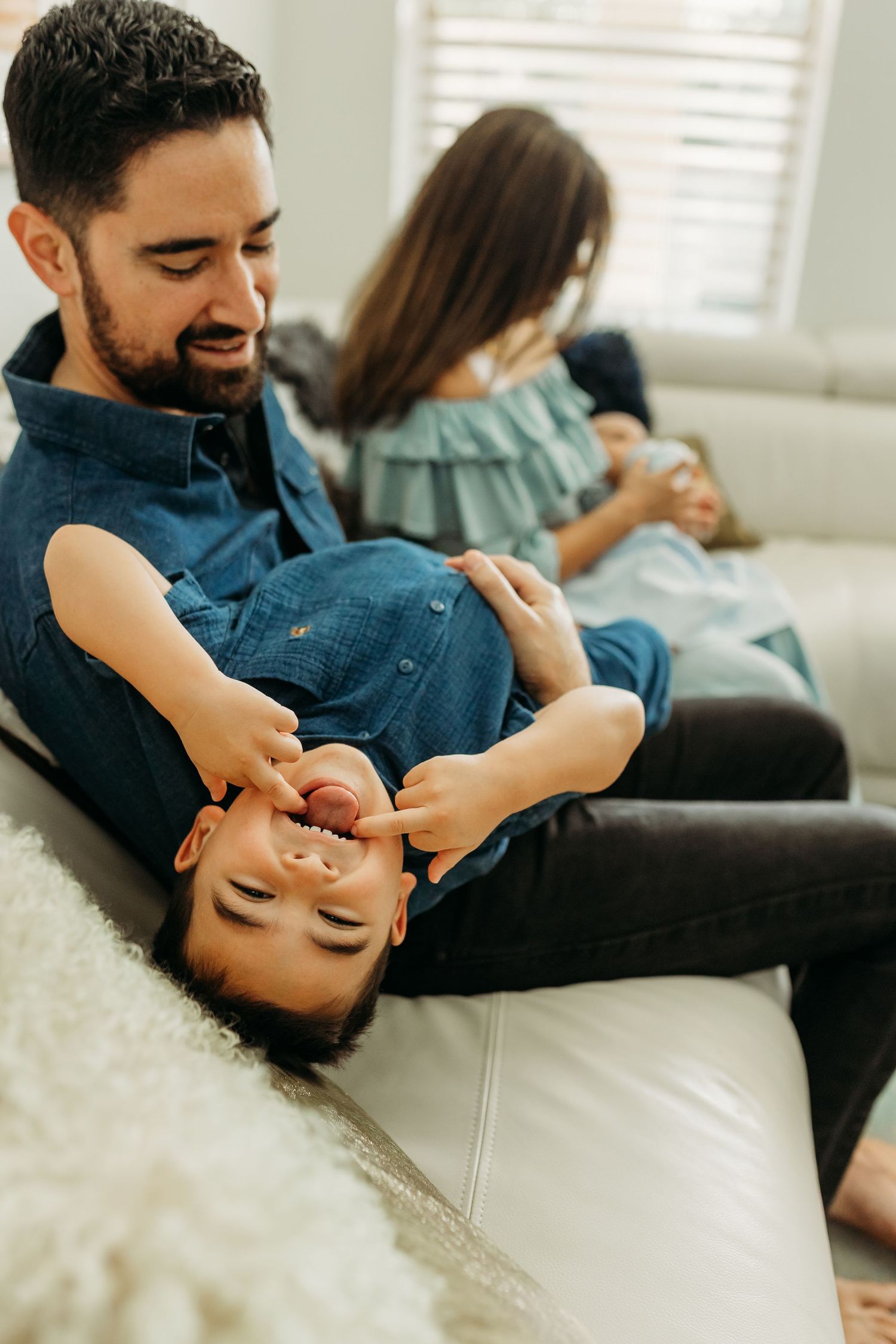 Father playfully holding young son upside down on white leather couch while family relaxes nearby.