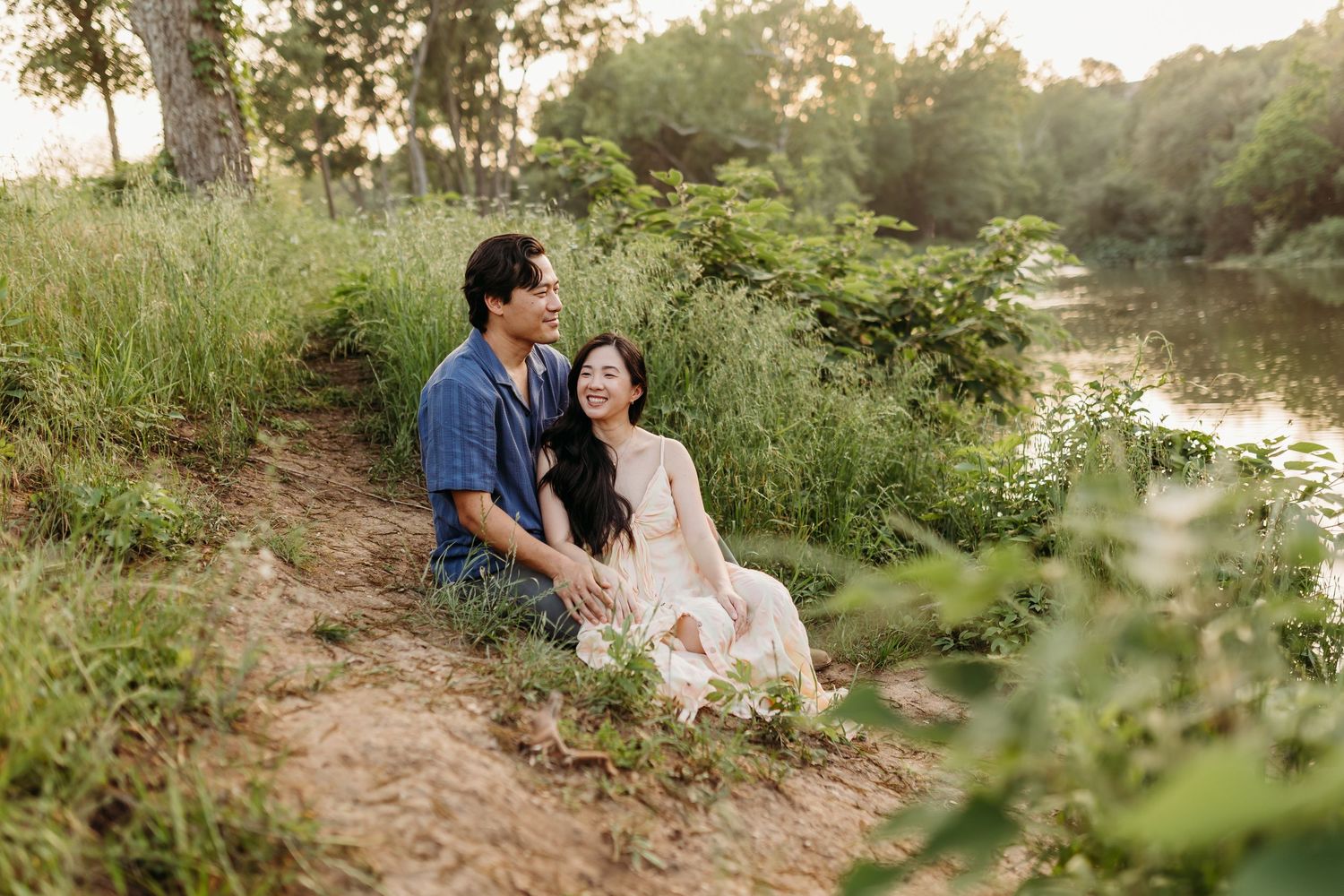 A smiling couple sits together on a sandy riverbank surrounded by green foliage in soft sunset light.