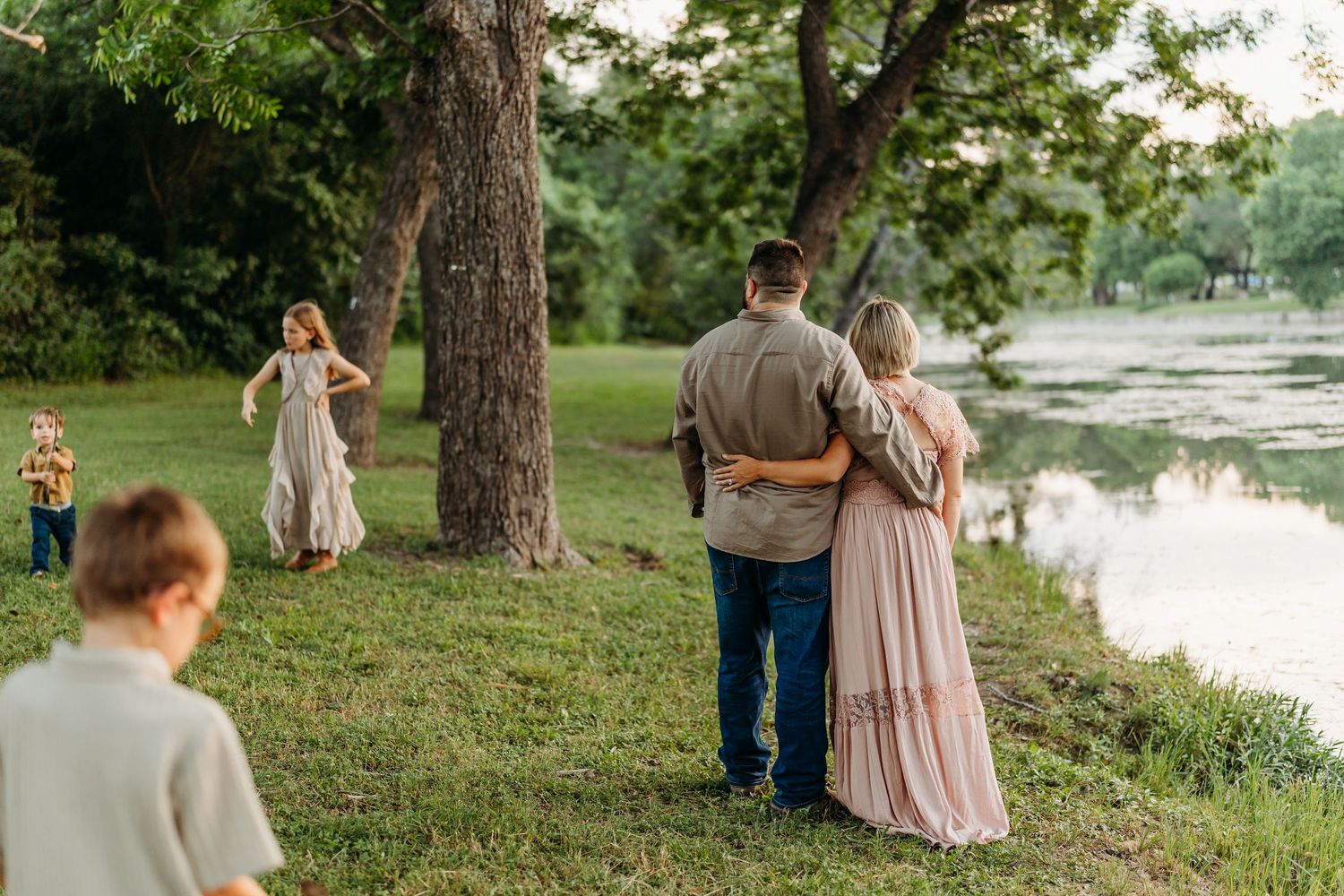 Couple walking arm in arm toward a scenic lake while children play among tall trees nearby.