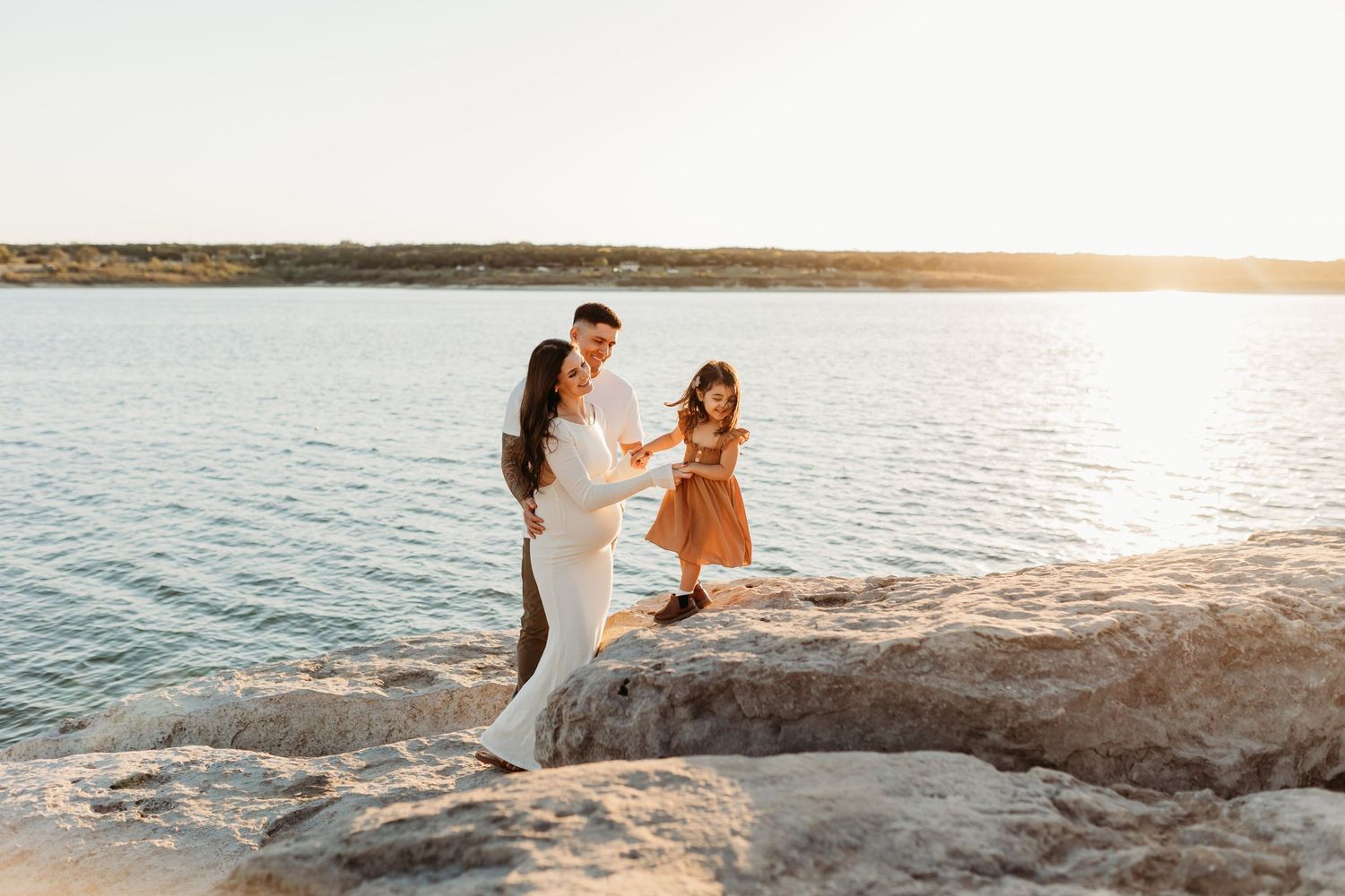 Pregnant couple and toddler daughter playing on rocky lakeside shore during a beautiful golden sunset.