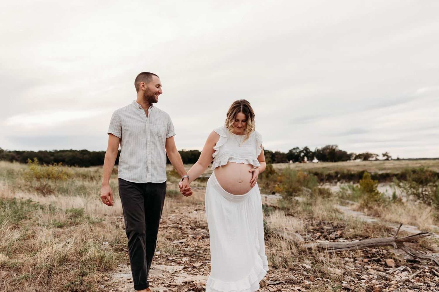 Pregnant couple holding hands walking through a field, man in gray shirt and woman in white maternity dress.