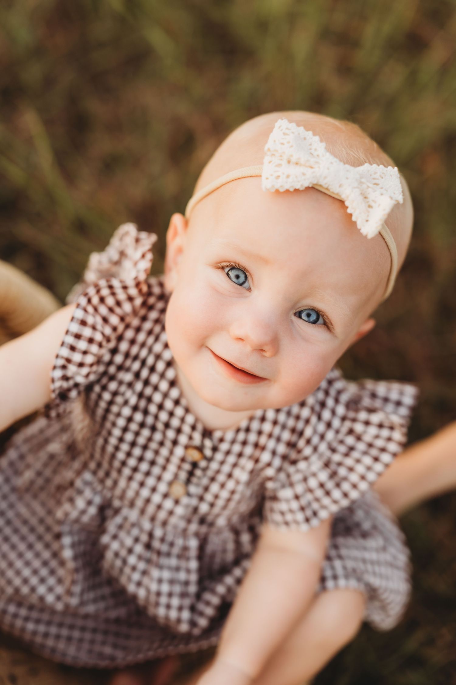 Adorable blue-eyed baby girl in gingham dress with white bow headband smiling outdoors.