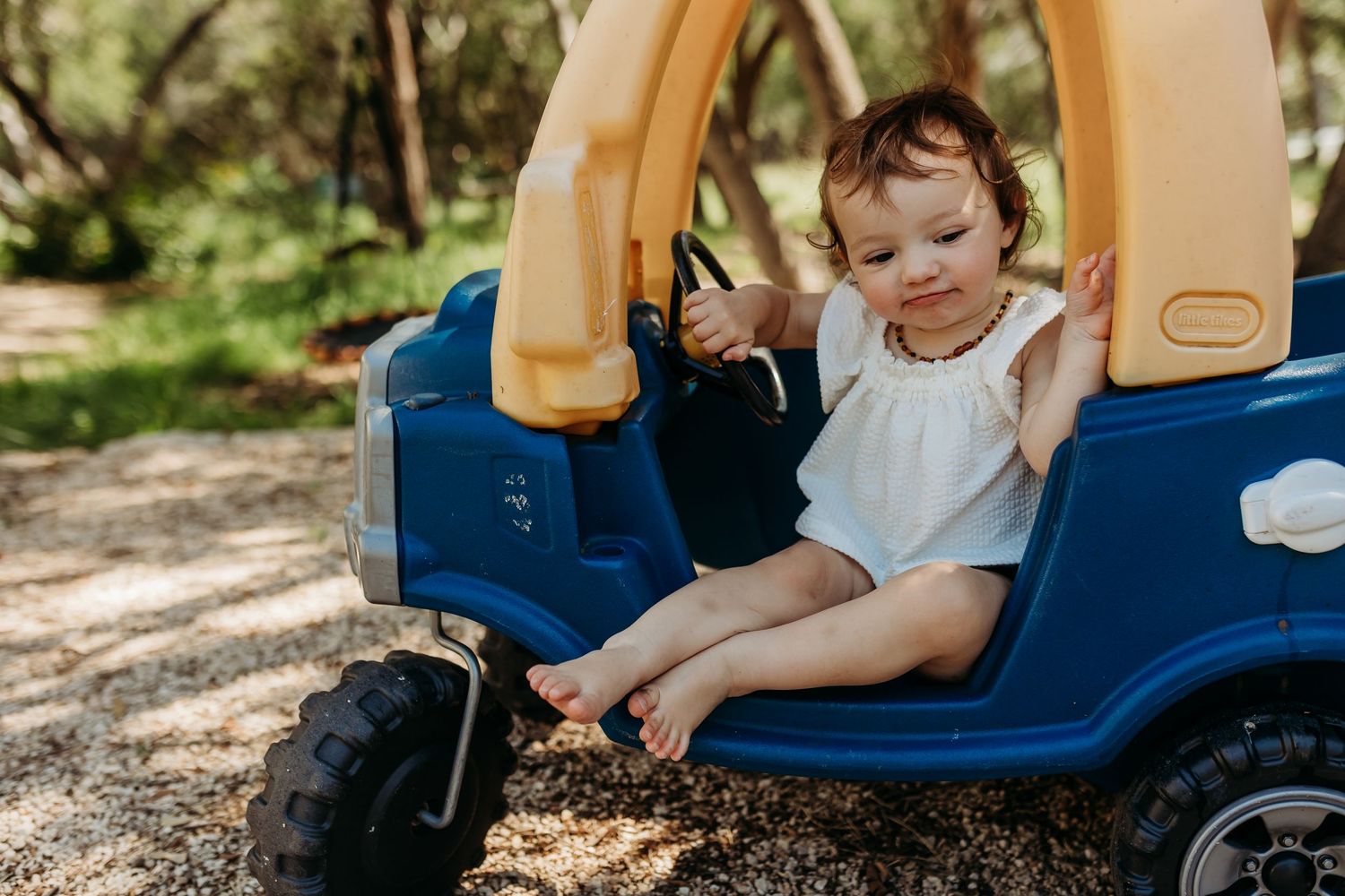 A cute toddler in a white dress sits in a blue plastic toy car on a shaded outdoor path.