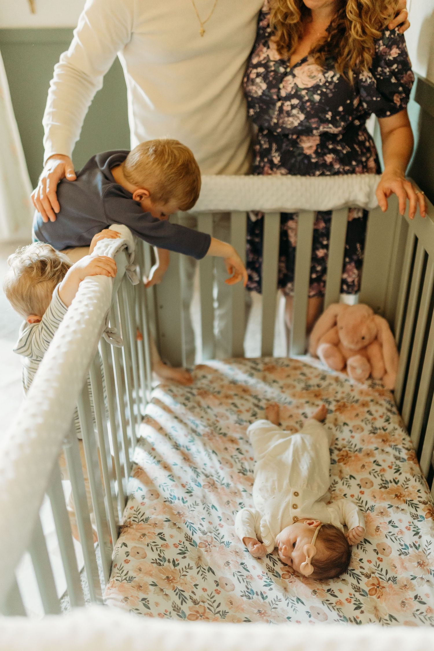 Parents watch over toddler climbing crib rail to peek at newborn baby lying inside.