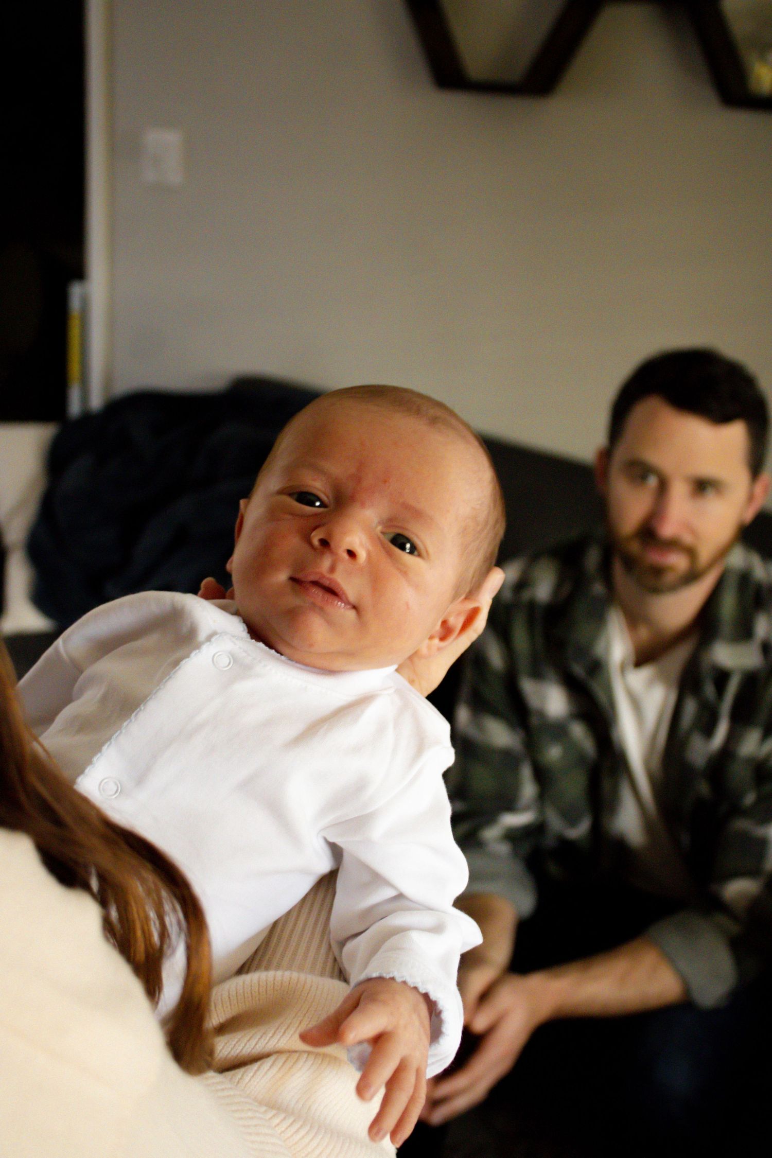mom holding baby, dad in background looking at baby boy, baby boy looking at camera