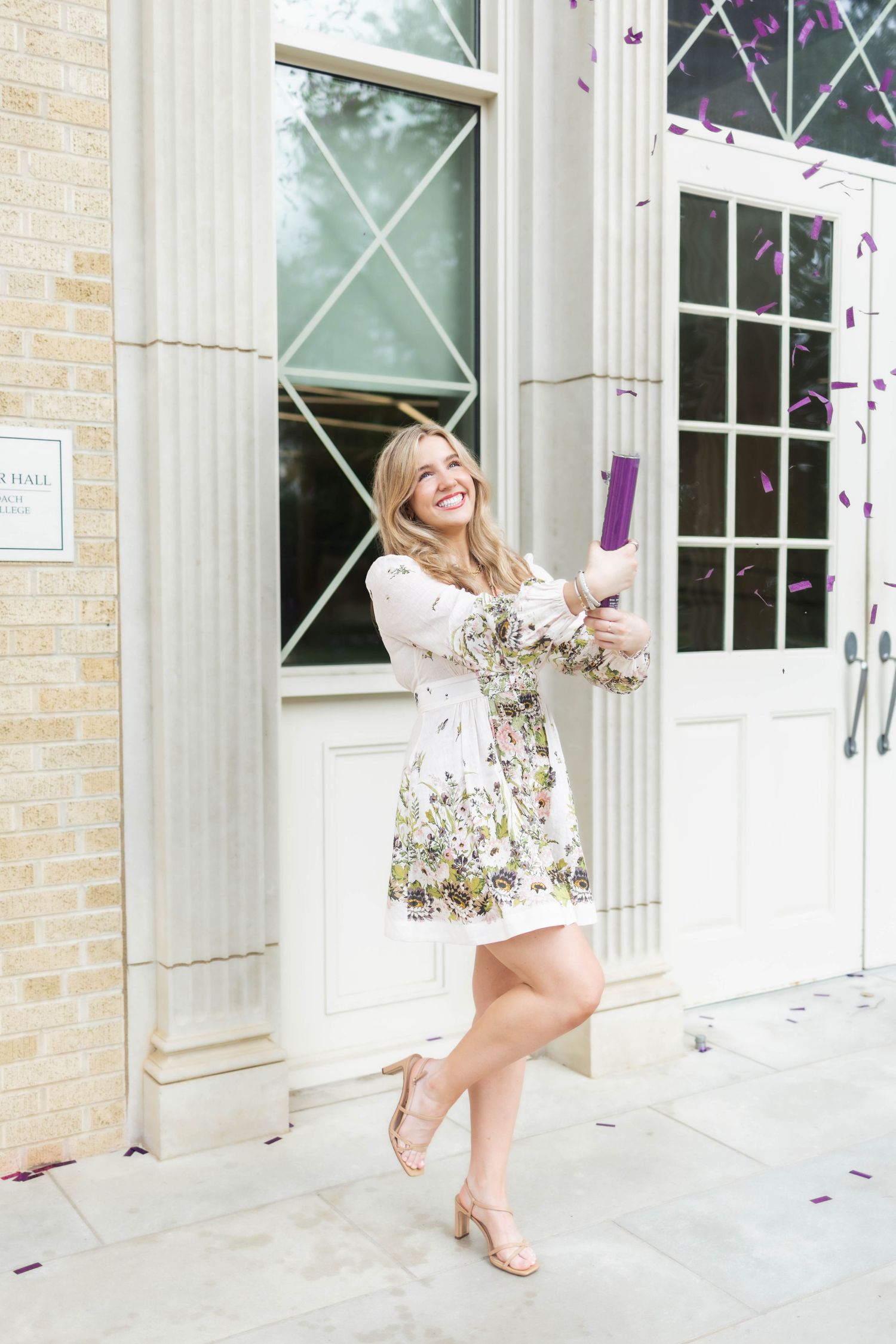Smiling blonde woman in floral dress celebrating with confetti popper outside elegant building columns.