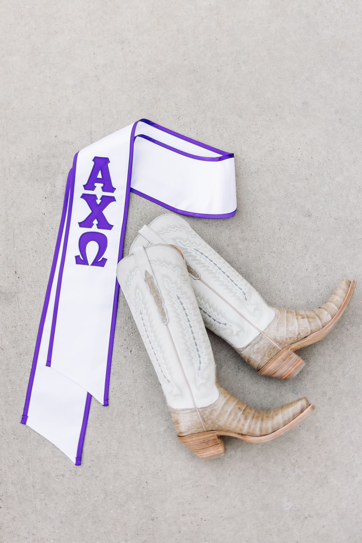 Alpha Chi Omega sorority sash with purple Greek letters displayed beside white cowboy boots on gray concrete.