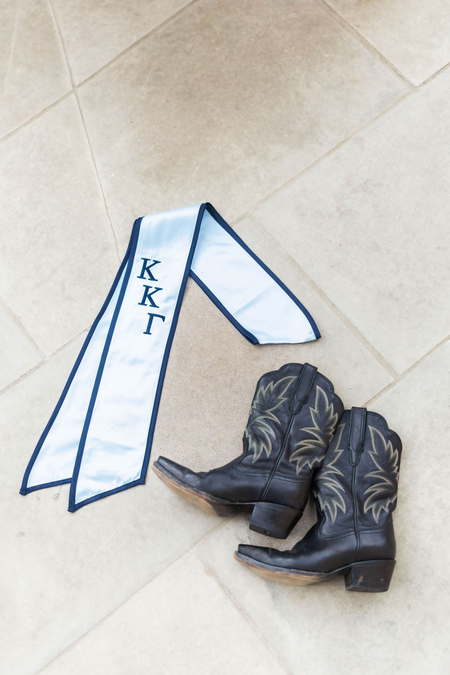 Kappa Kappa Gamma sorority graduation sash with navy trim beside black leather cowboy boots on tile floor.