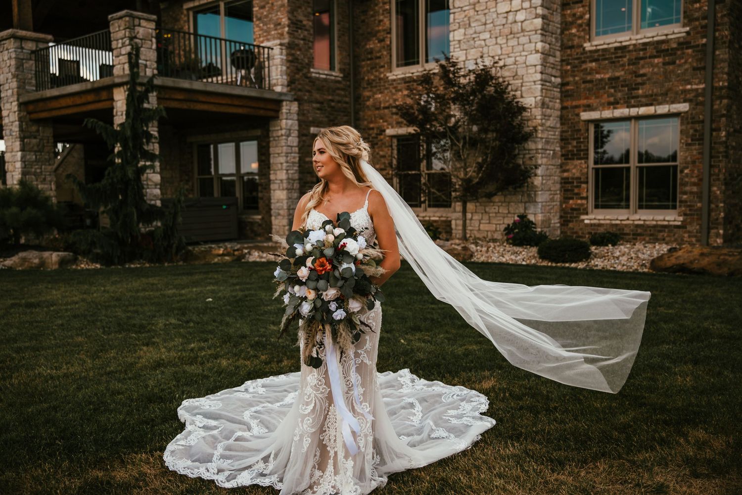 Bride in fitted lace gown with flowing veil stands on lawn near brick building.