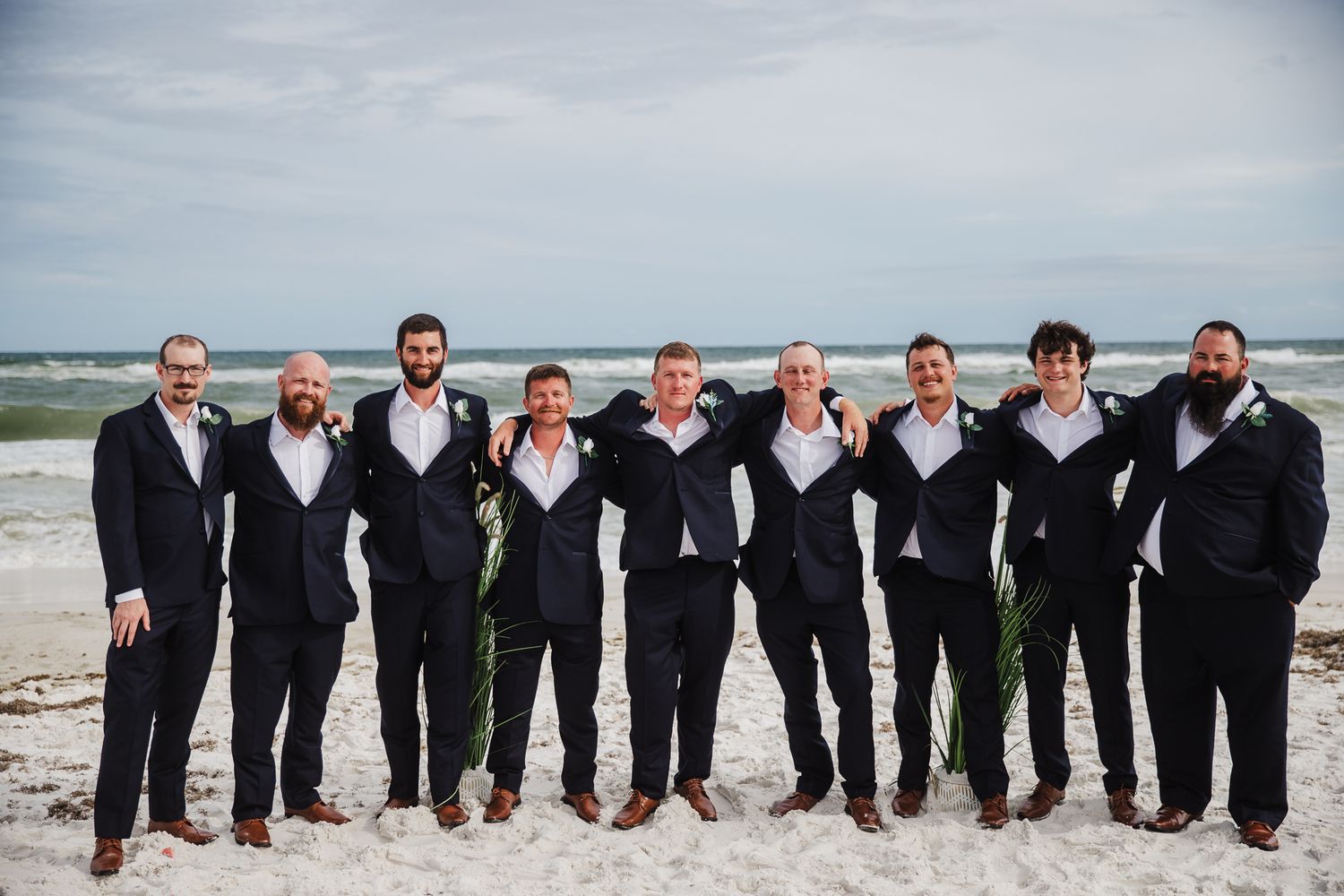 A group of nine groomsmen in black suits standing together on a beach with the ocean waves in the background.