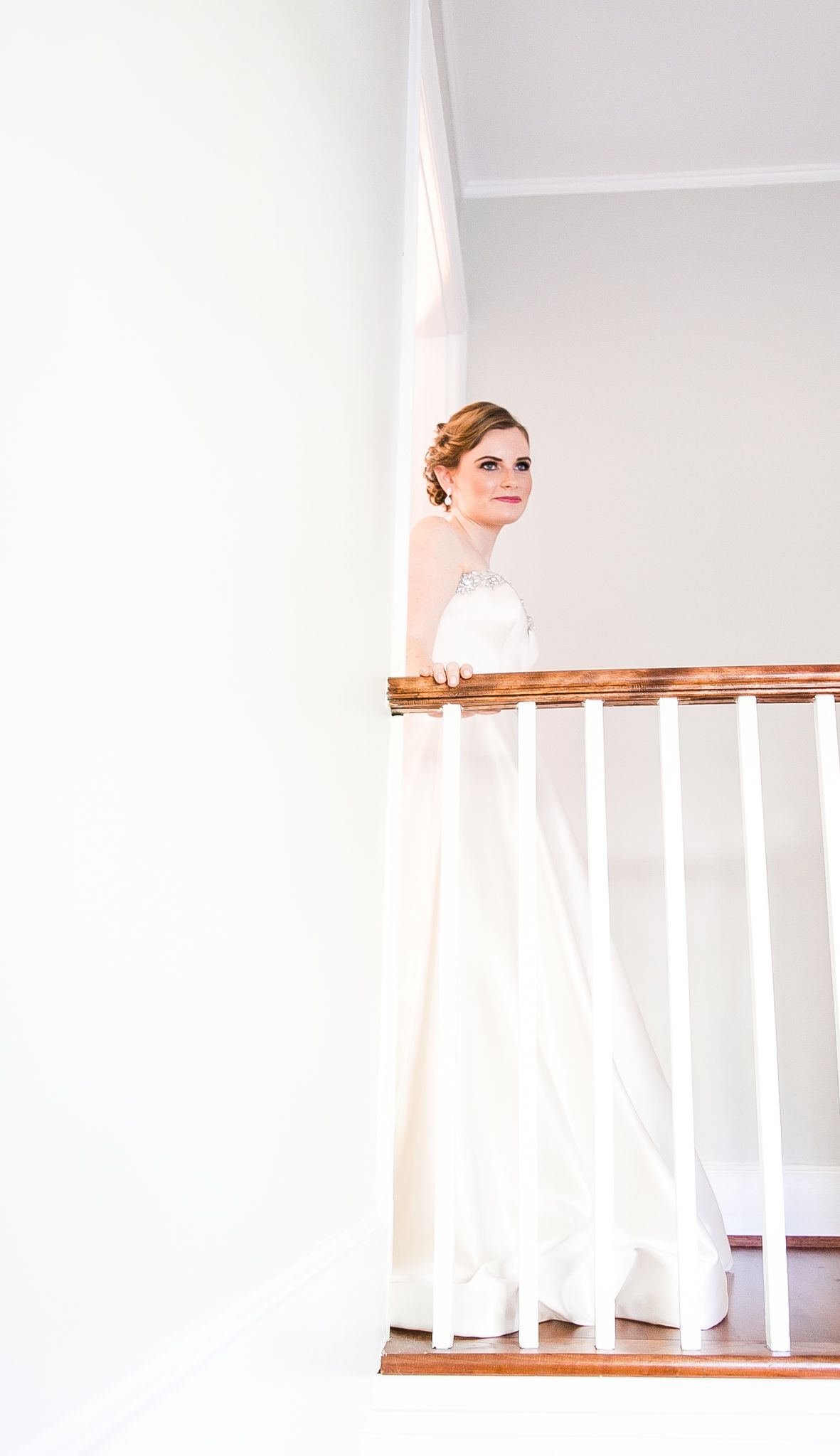 A bride in a white wedding gown stands elegantly on a staircase landing with white walls and wooden railings.