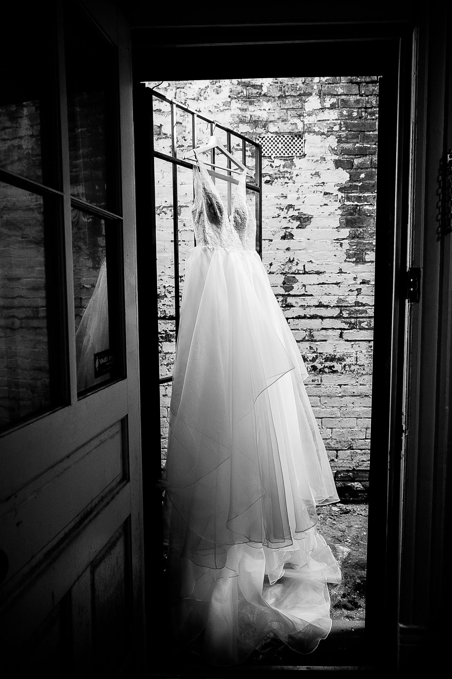 A white wedding dress hangs elegantly in a doorway against a weathered brick wall in black and white photography.