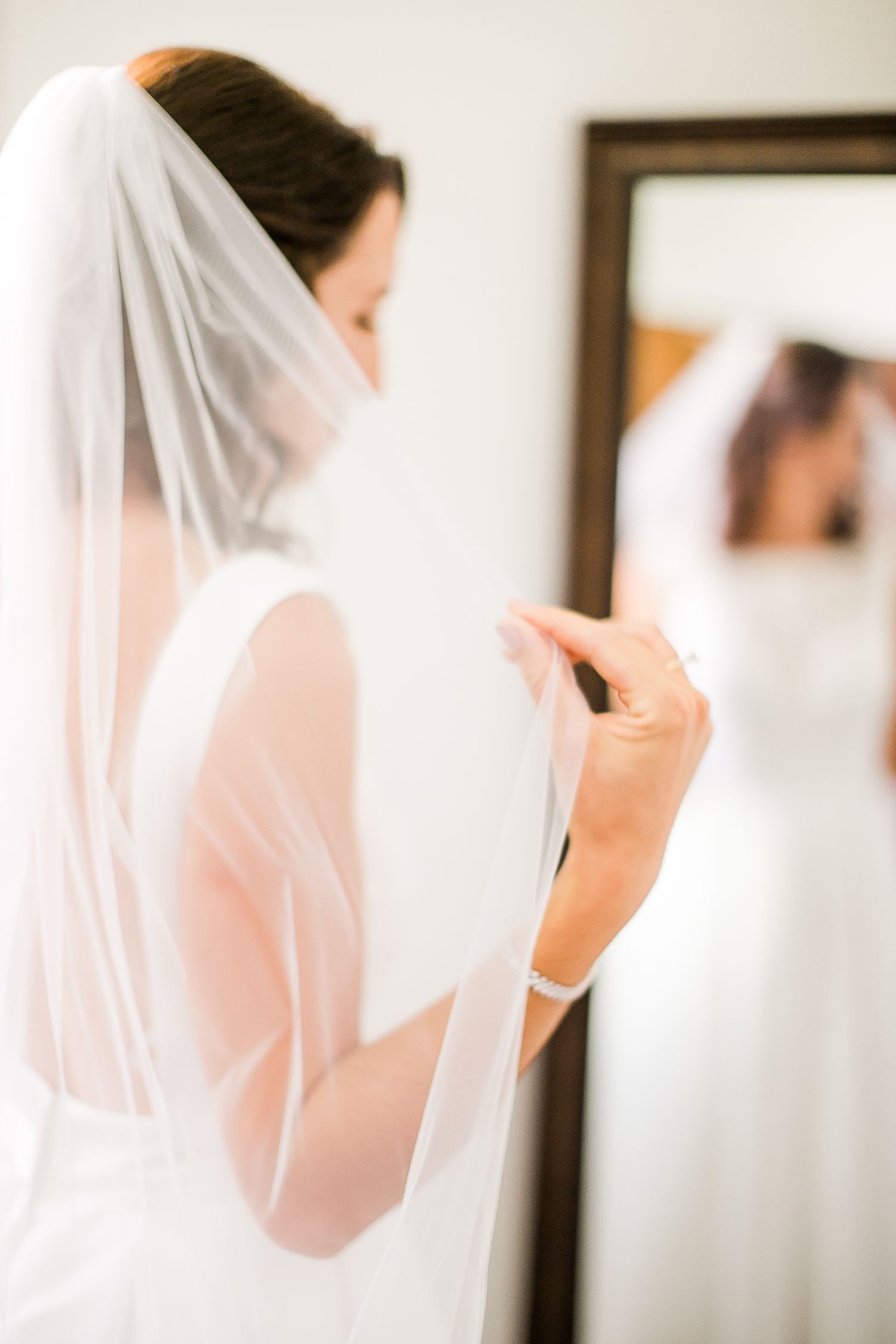 A bride adjusts her delicate white veil while looking in a mirror during wedding preparations.