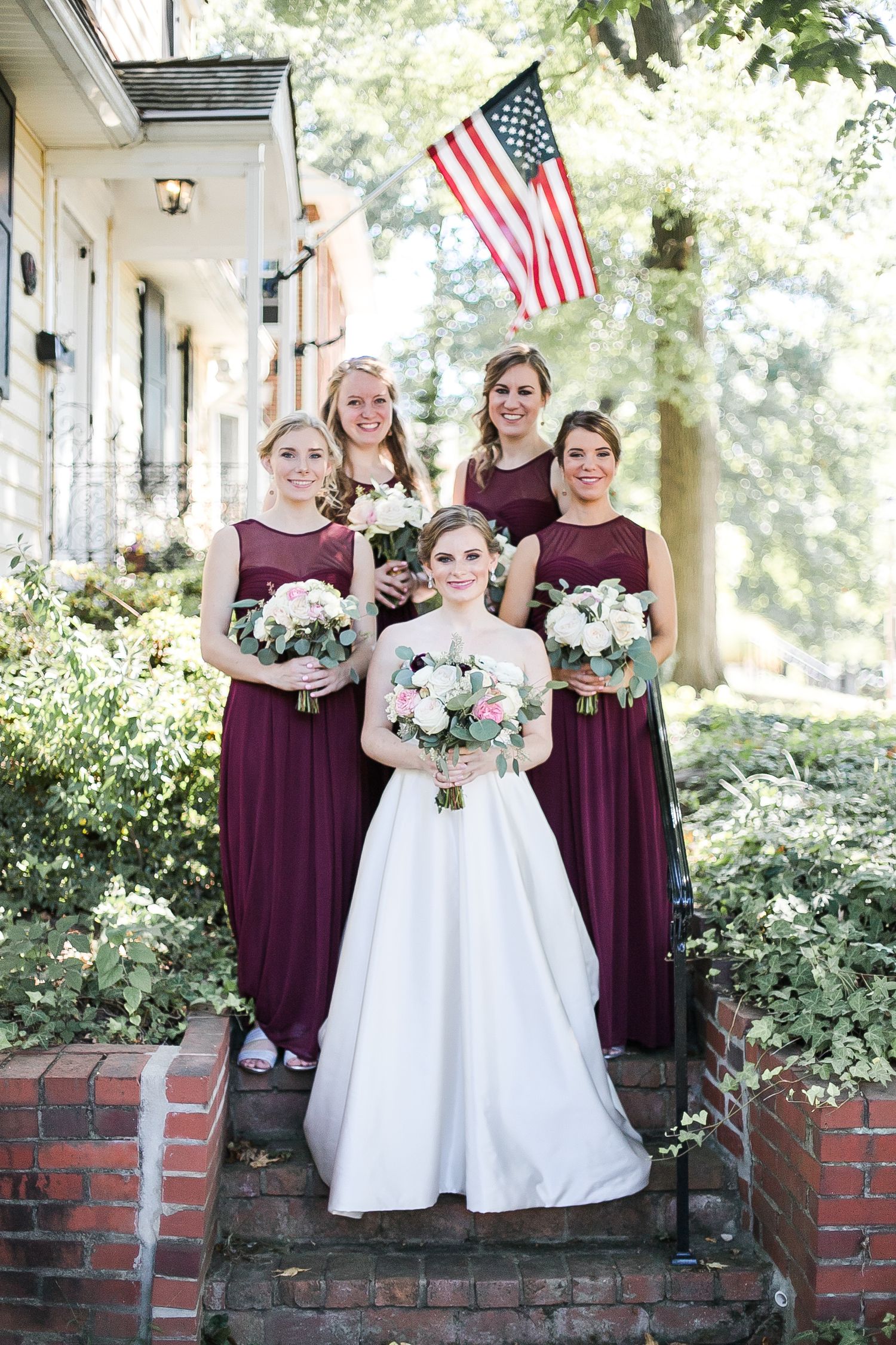 Bridal party in burgundy bridesmaid dresses and white wedding gown pose on brick steps under the American flag.