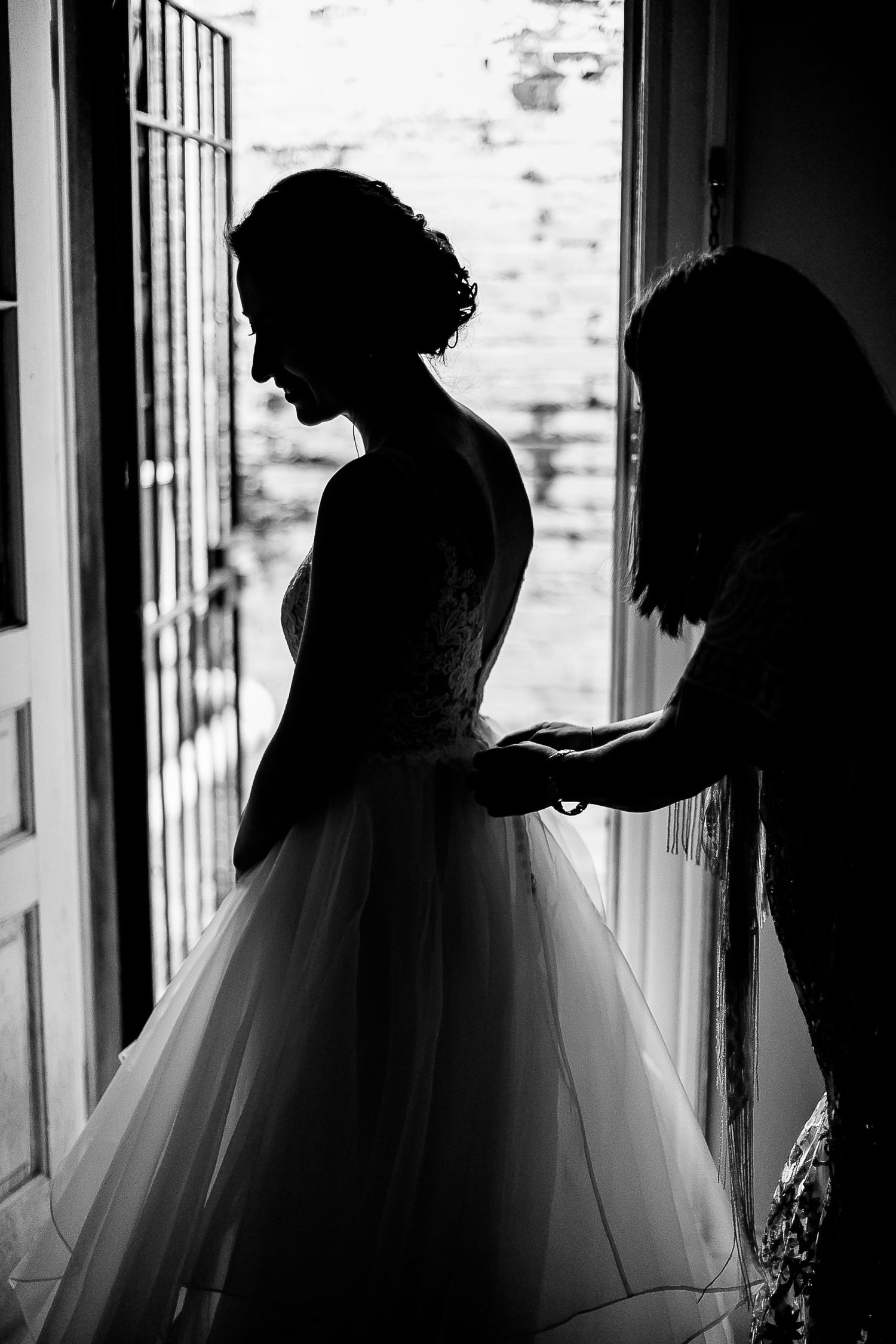 A black and white silhouette of a bride in wedding dress getting ready by a window with soft natural light.