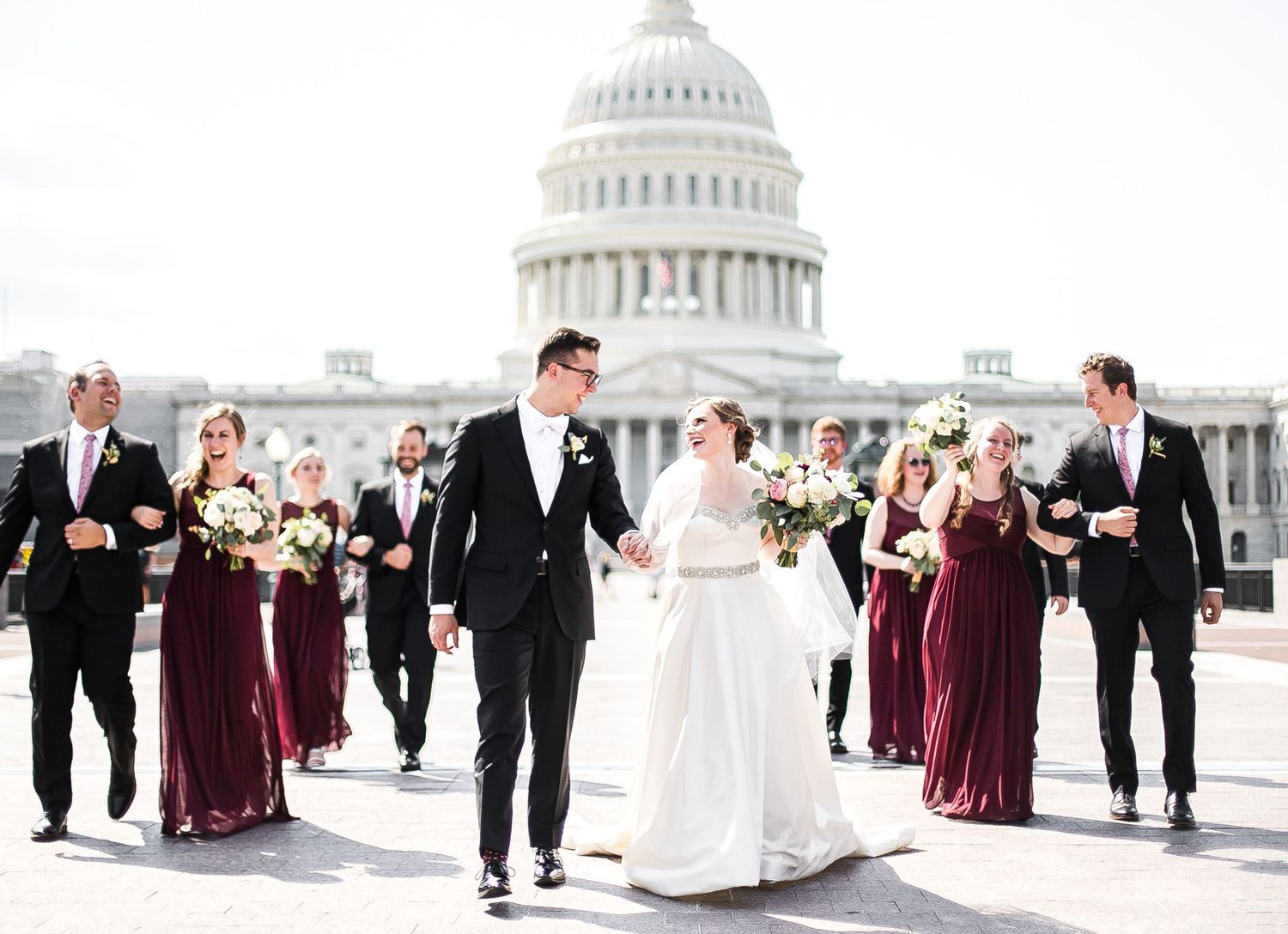 The wedding party walks in front of the US Capitol Building with bridesmaids in burgundy dresses on a bright day.