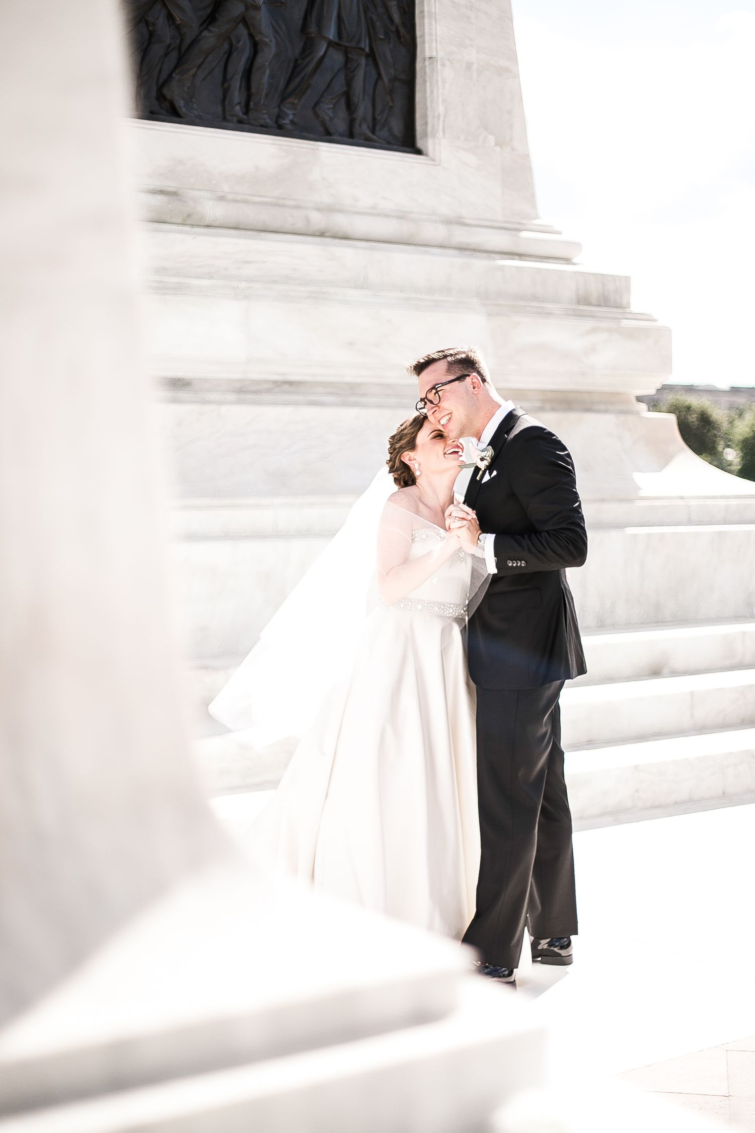 A romantic wedding couple embraces on white marble steps in bright sunlight.