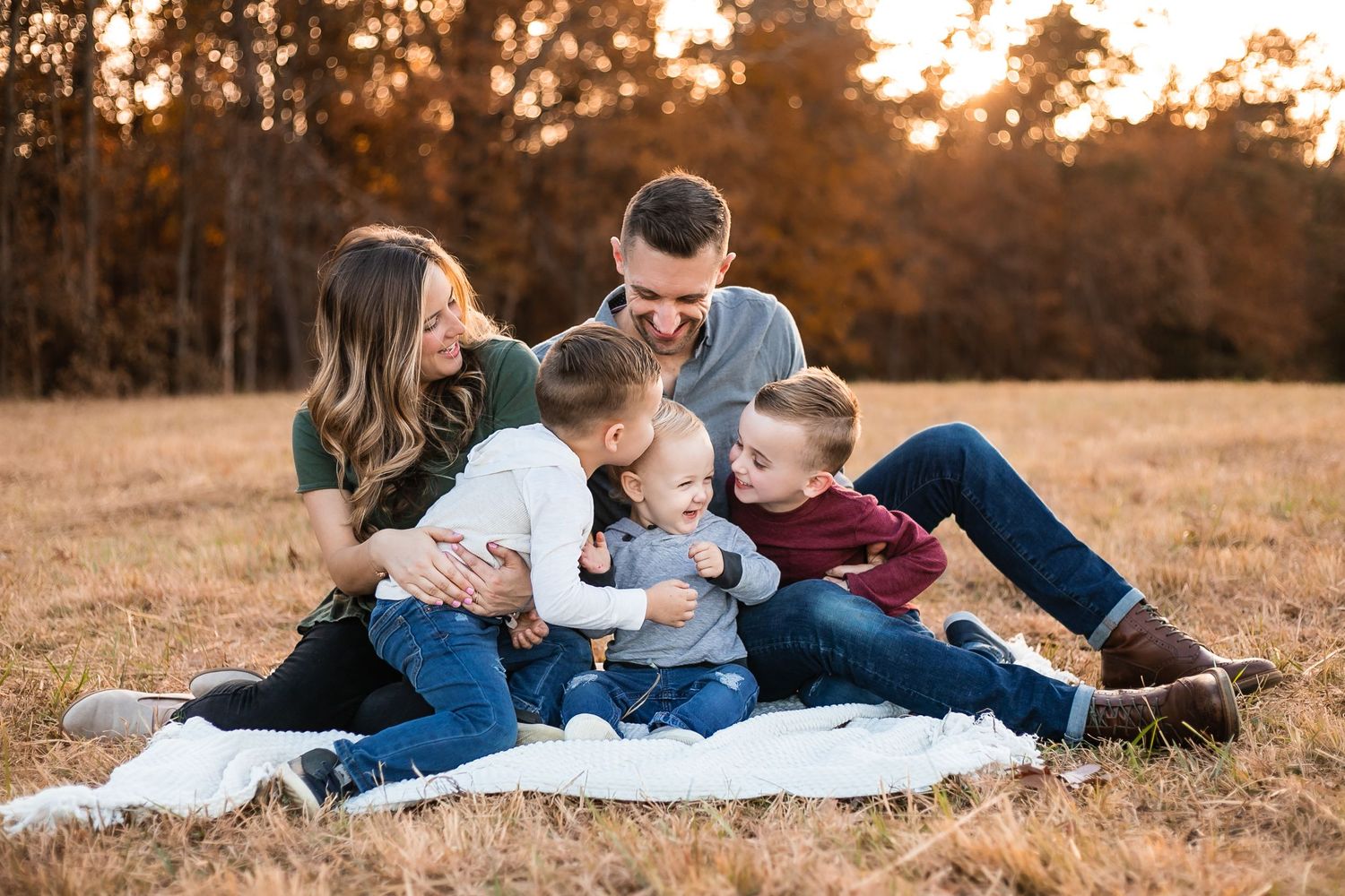 A family sits together on a blanket in a golden field at sunset, sharing tender moments.
