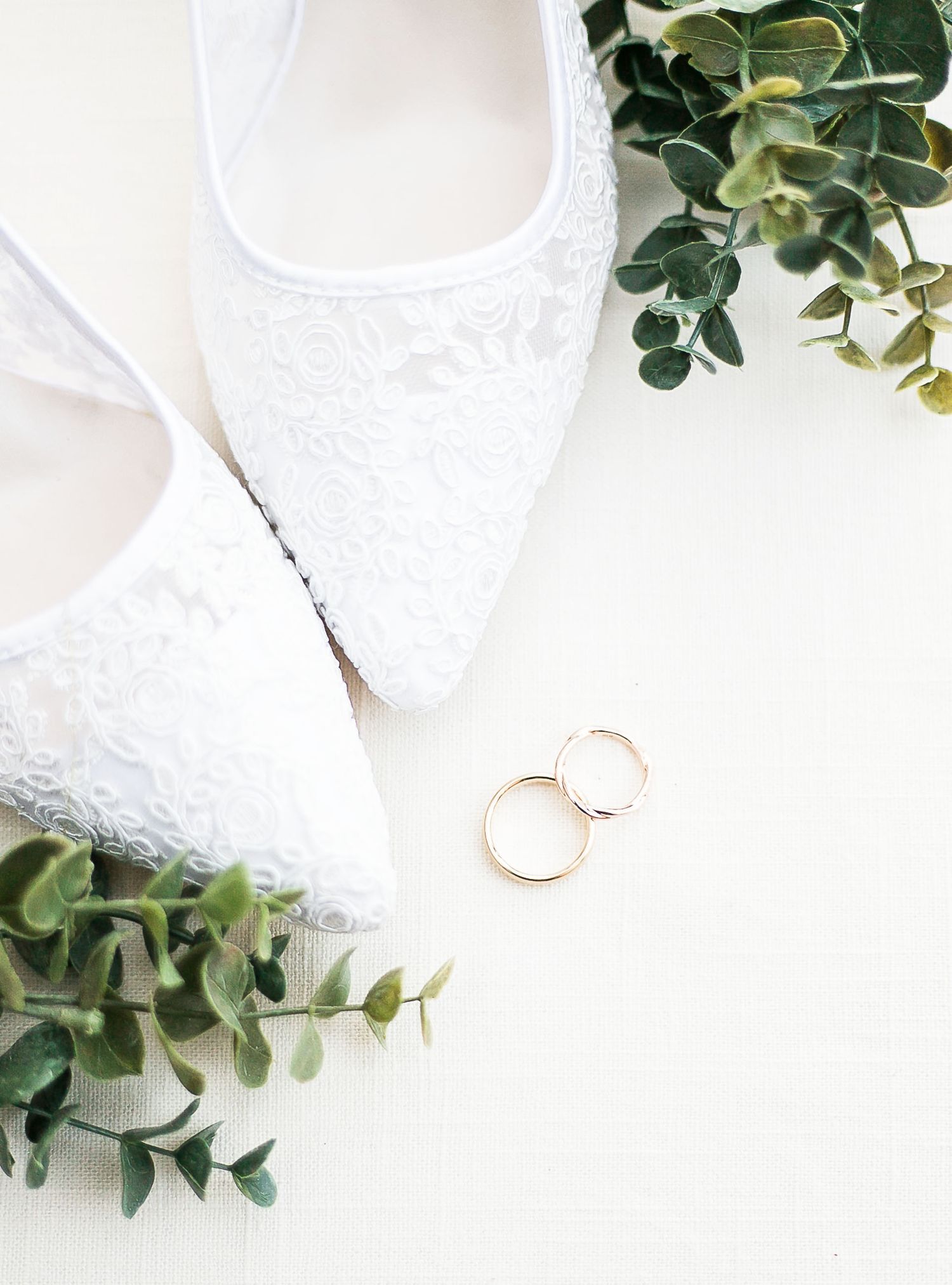 White lace wedding flats with gold wedding rings and eucalyptus leaves on white background.