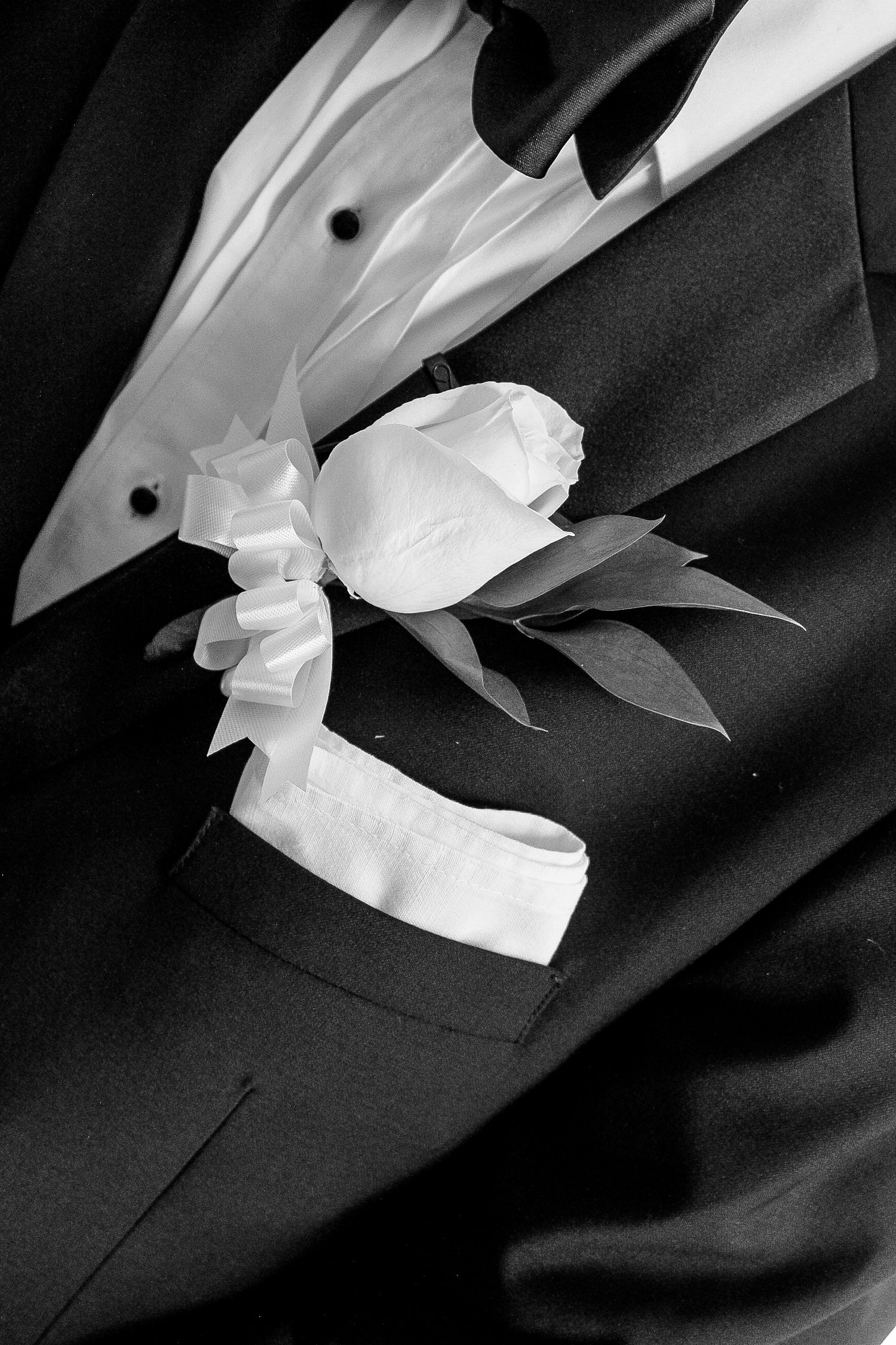 A white floral boutonniere pinned on a black tuxedo lapel in black and white photography.