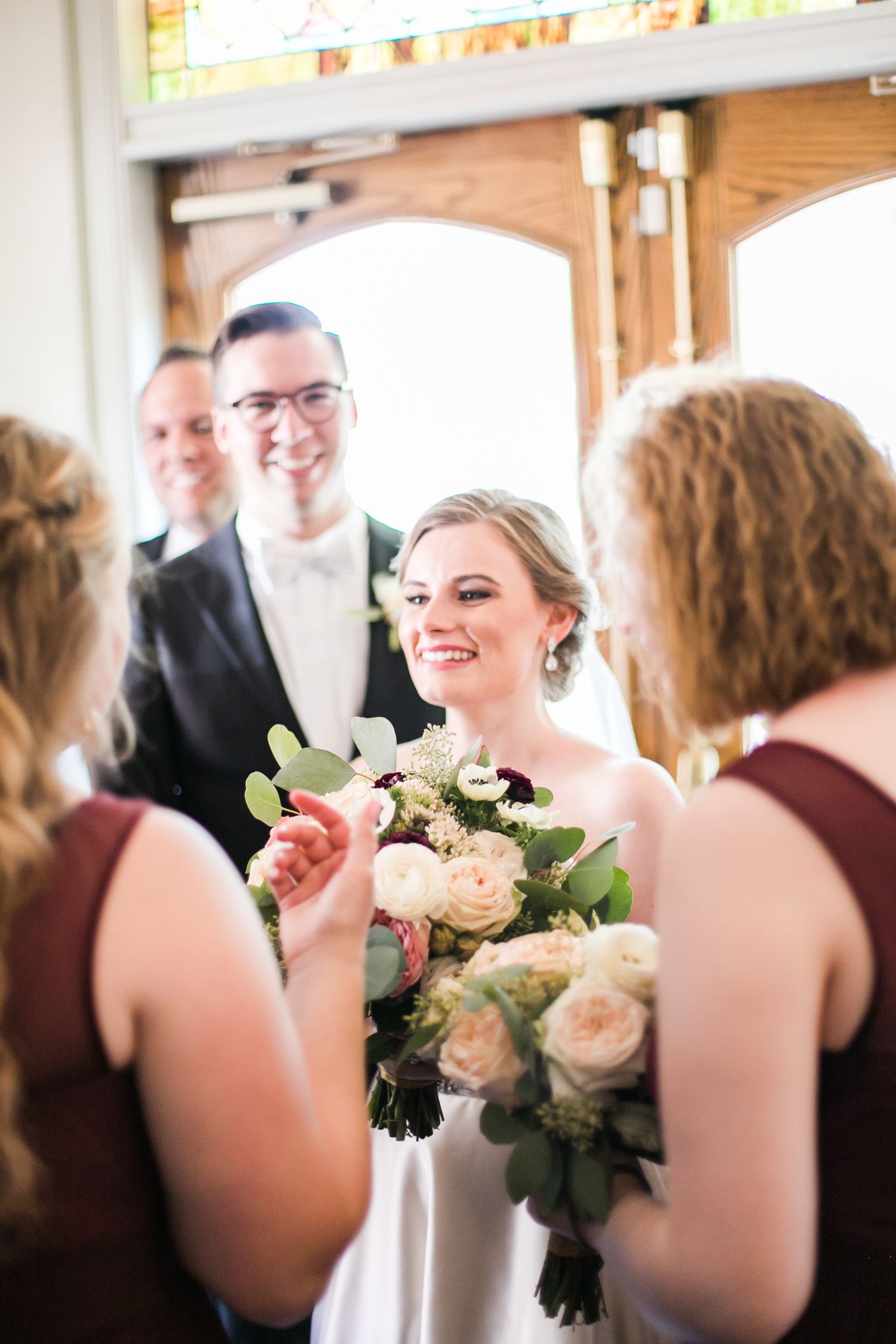 A couple together on their wedding day shares a moment indoors with bridesmaids in burgundy dresses and elegant floral bouquets.