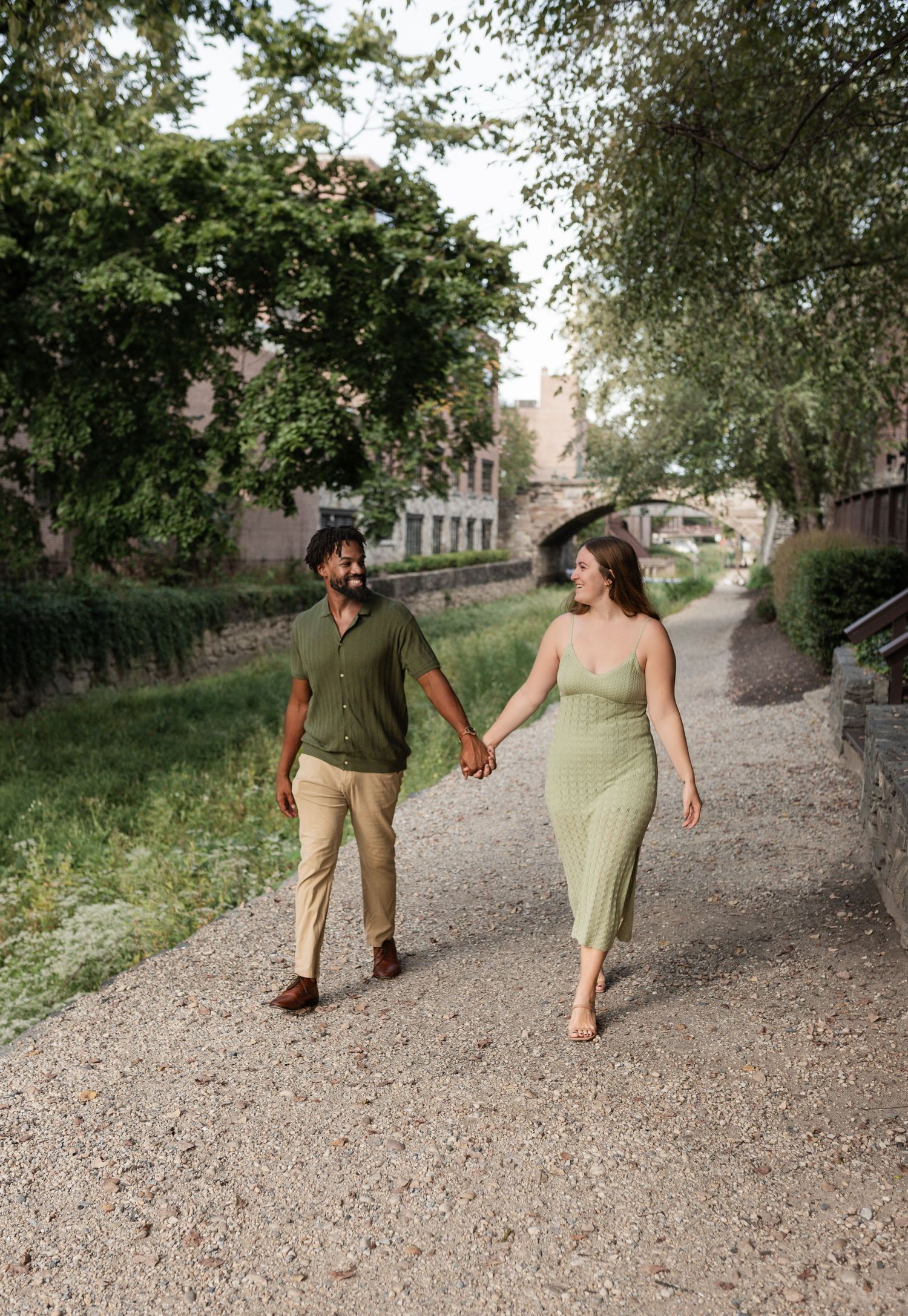 A couple walks hand in hand along a tree-lined gravel path on a pleasant summer day.