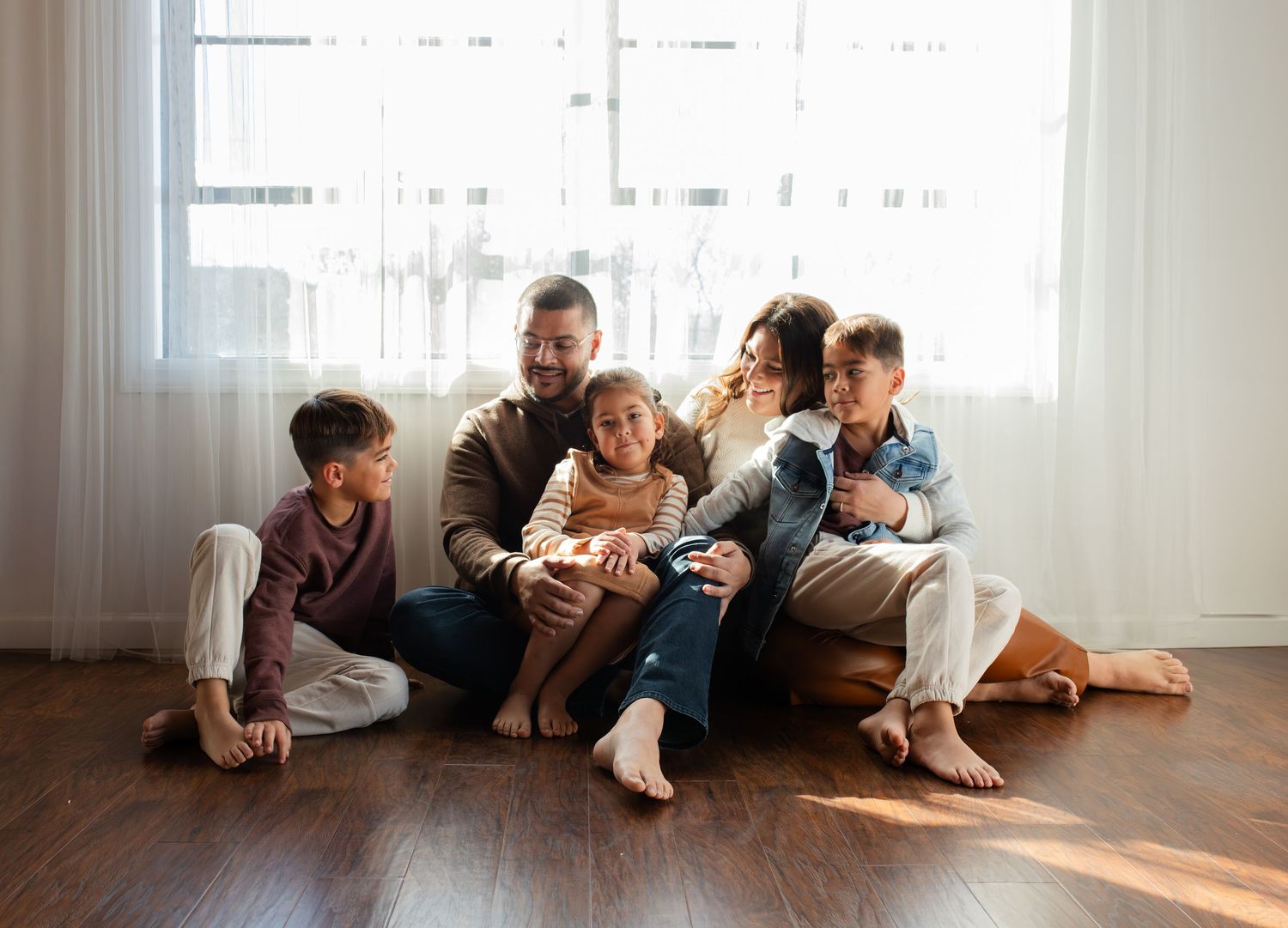 A family sits together on a wooden floor at home, laughing and enjoying quality time in front of sheer curtains.