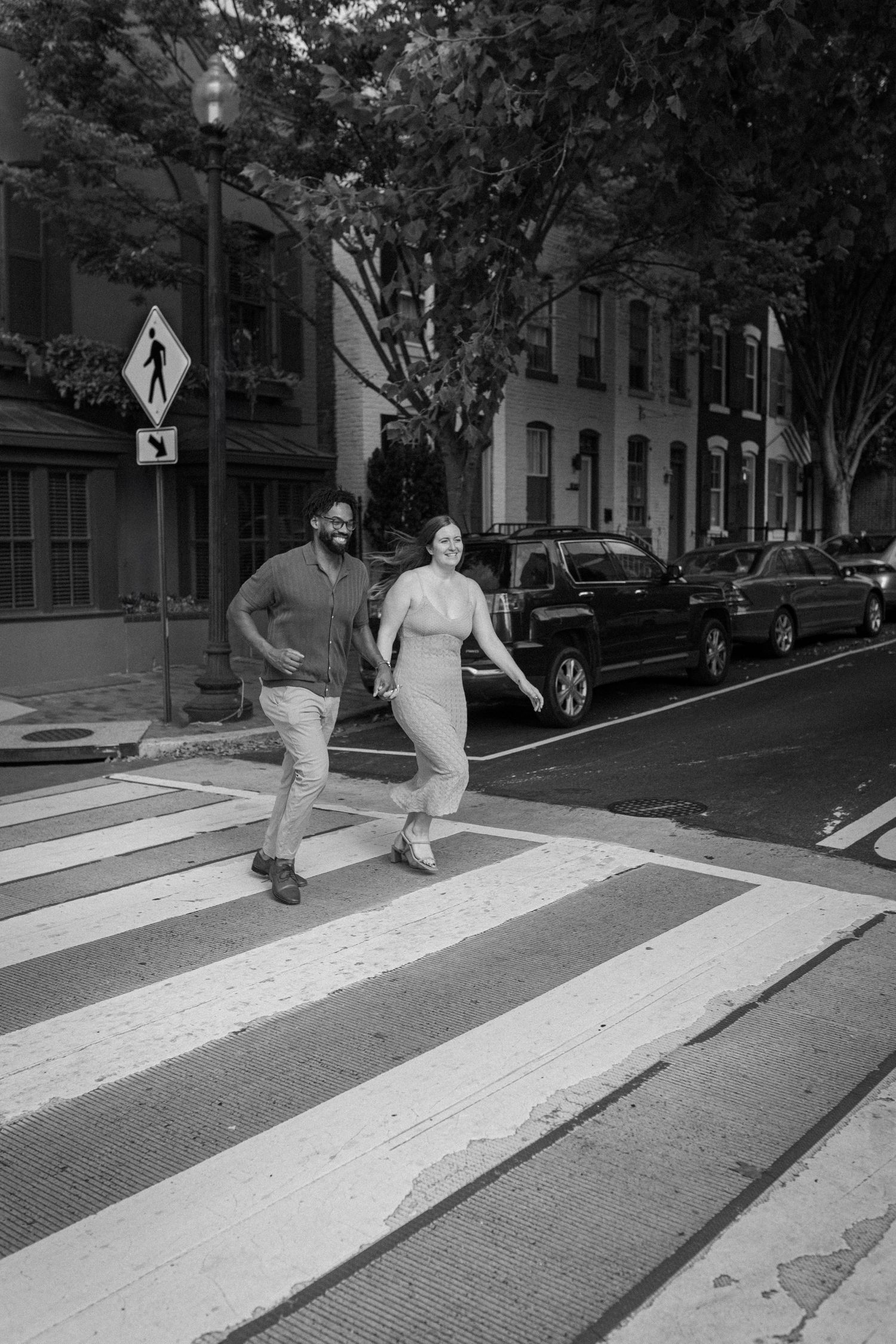 A couple crosses a crosswalk together on a tree-lined city street with parked cars and a pedestrian sign visible.