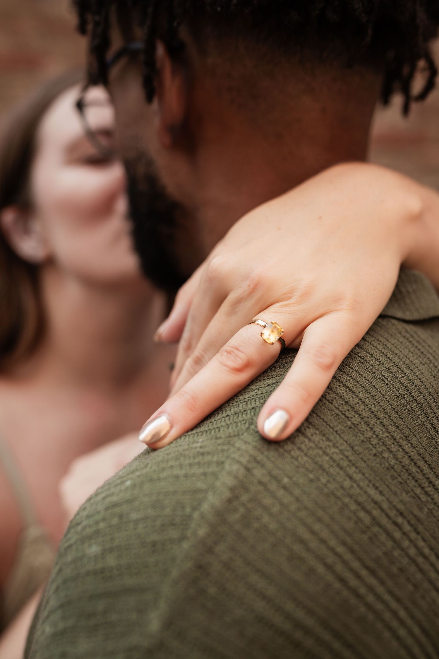 A woman's hand with nude nail polish rests on a green knit sweater showing an engagement ring.