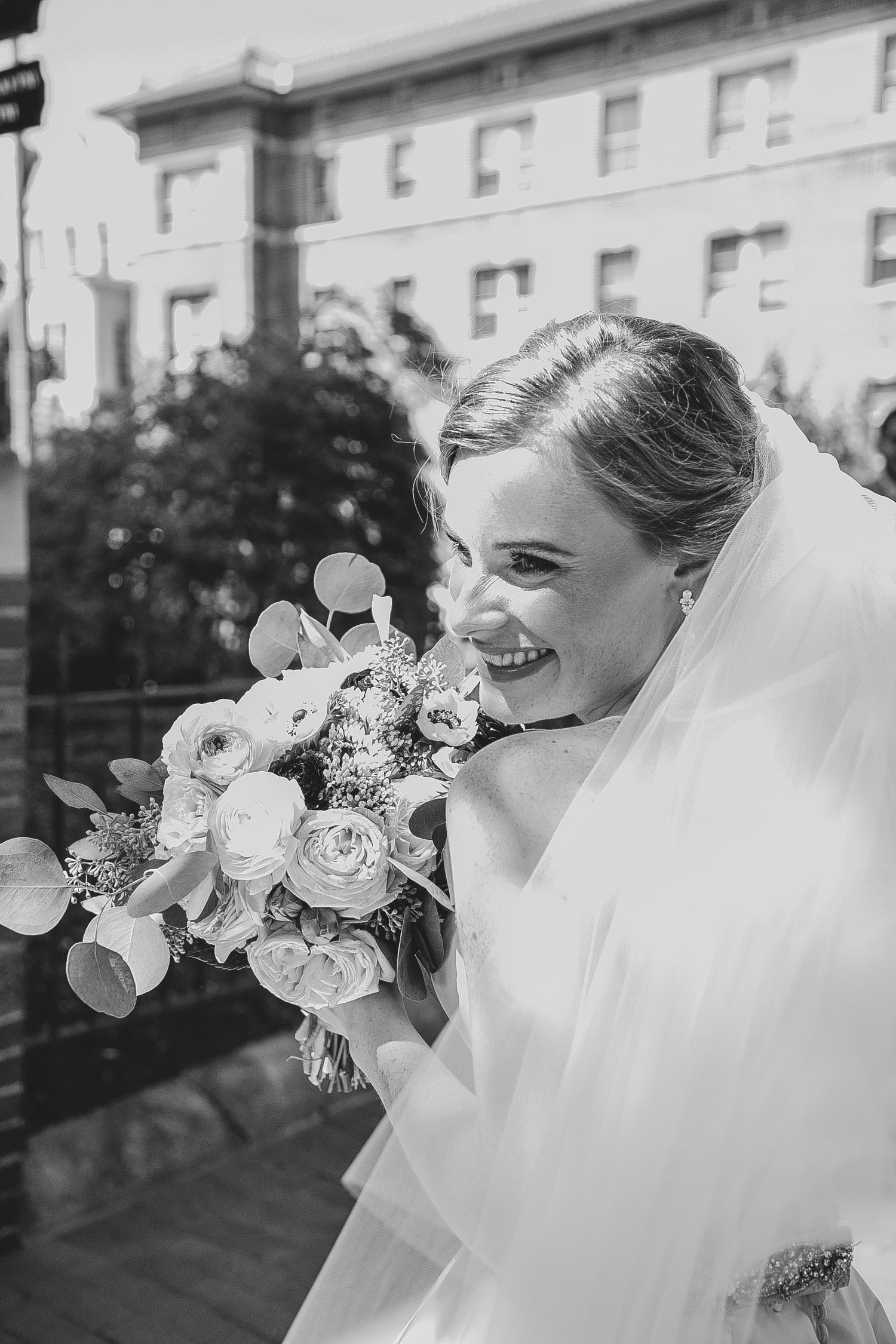 A joyful bride in a white veil smiles while holding an elegant bouquet of roses and eucalyptus against an urban backdrop.